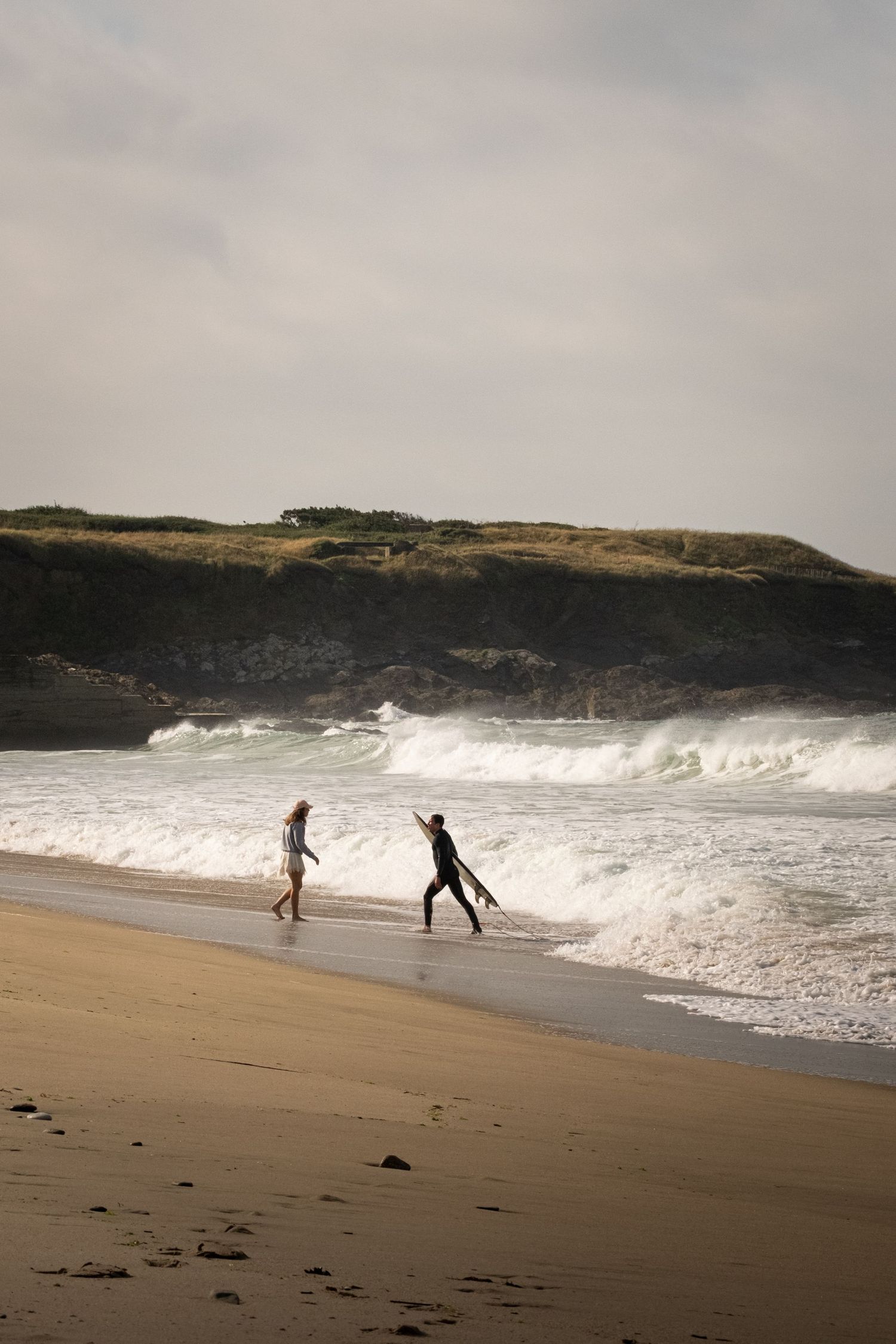 Two surfers walk along a sandy beach with crashing waves and rocky cliffs in the background on a cloudy day.