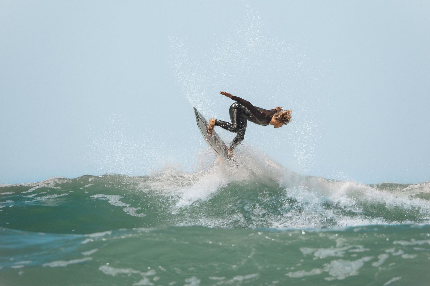 Surfer performing an aerial maneuver on a wave in the ocean wearing a dark wetsuit.