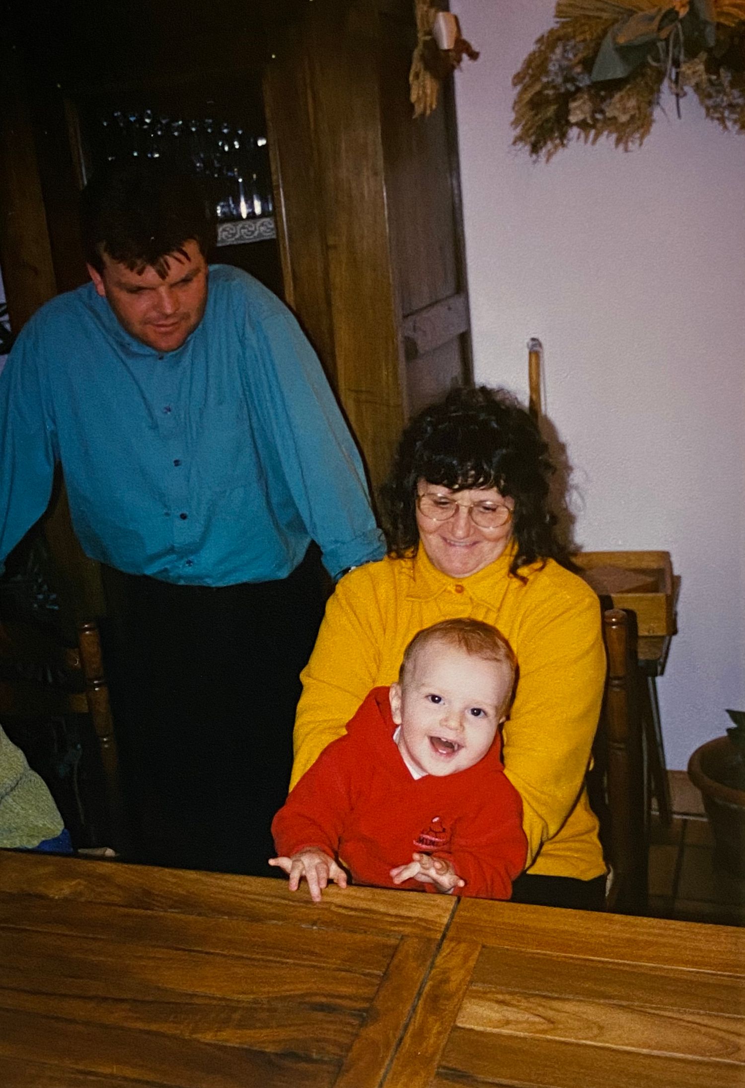 A smiling baby in red sits at a wooden table with two adults in a warm home setting.