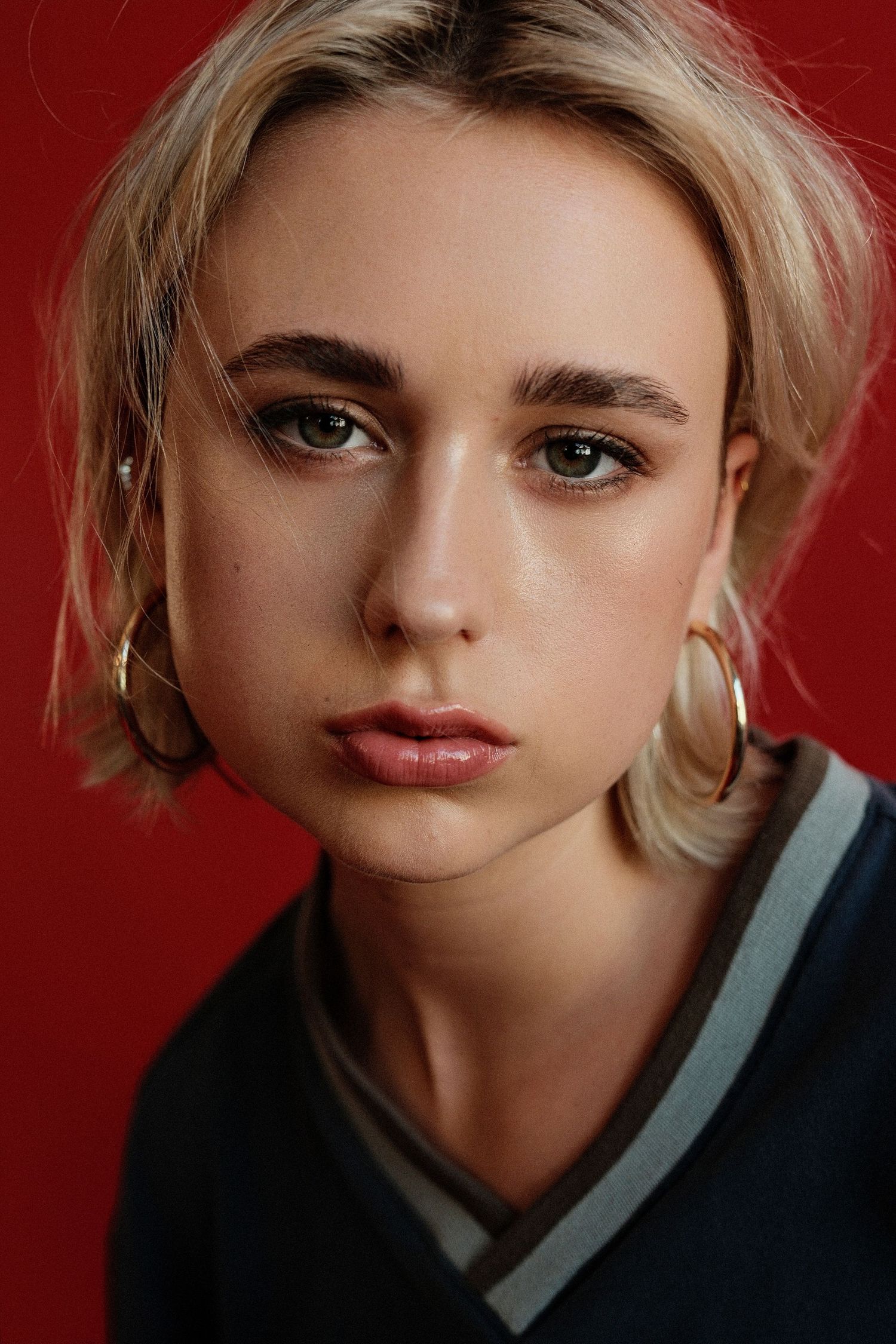 Young blonde woman with hoop earrings and short hair poses against a bold red background in a casual varsity-style top.