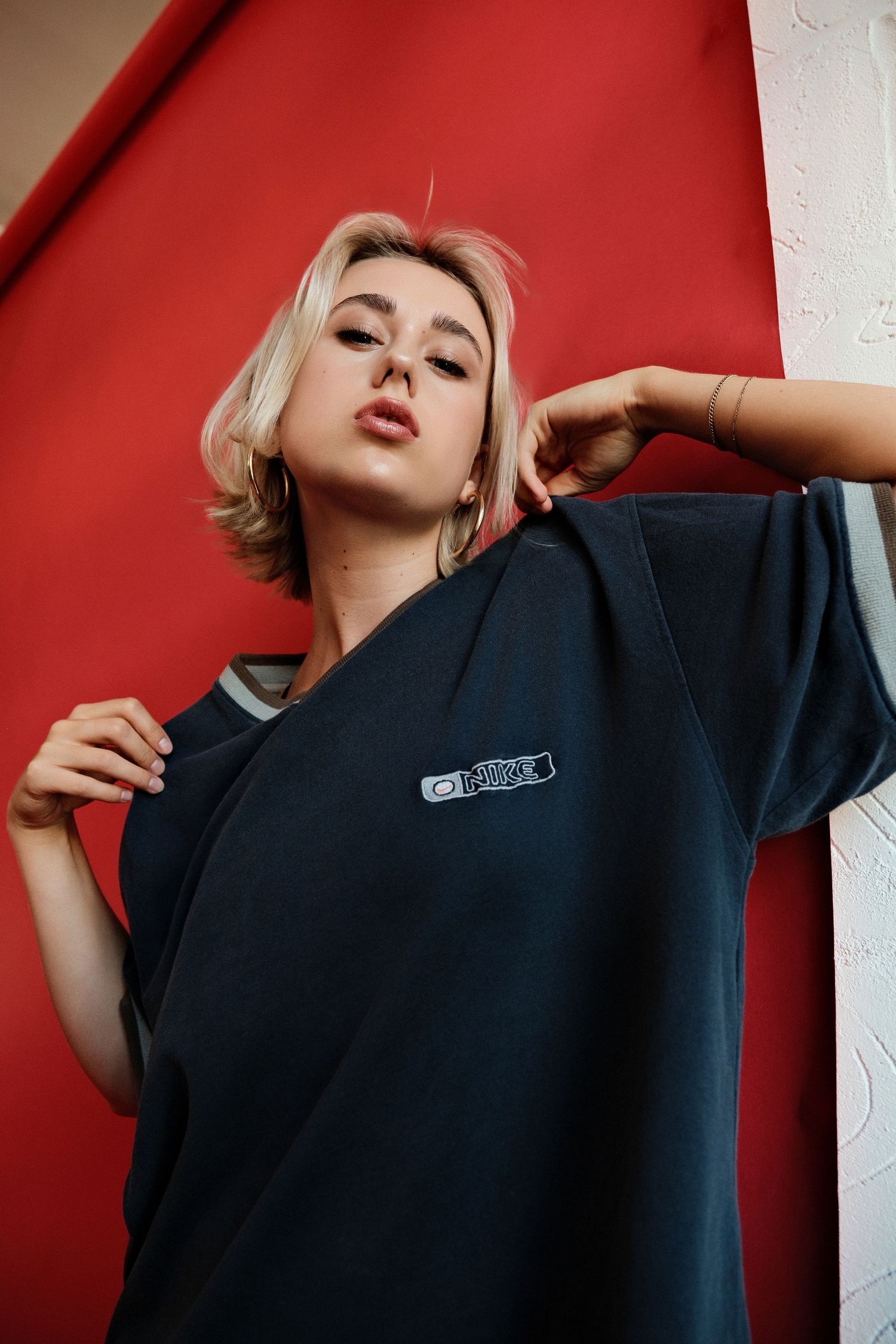 Young woman with blonde hair posing in an oversized dark navy t-shirt against a bold red and white background.