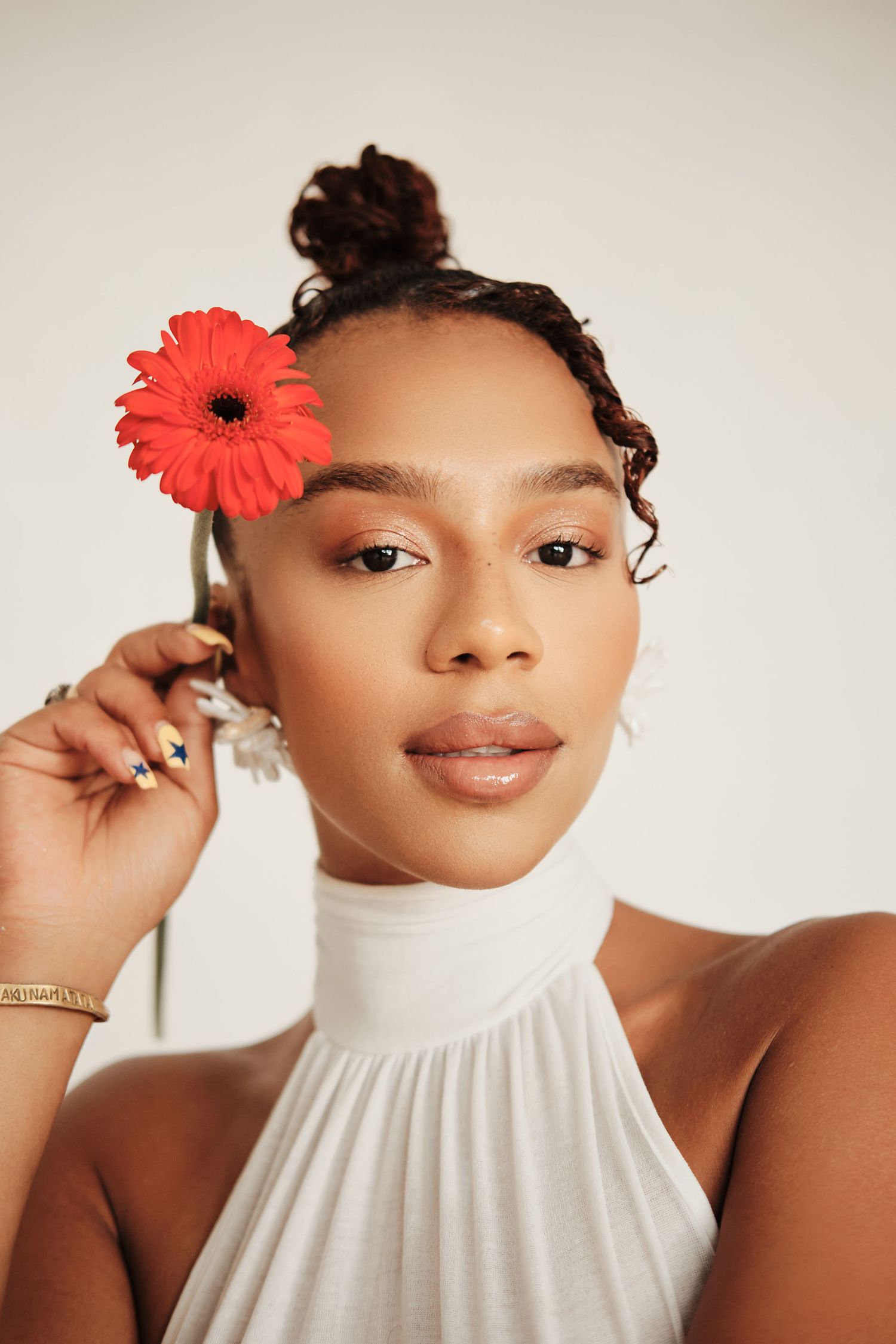 Young woman with curly updo holding a red gerbera daisy, wearing a white halter top against a light background.