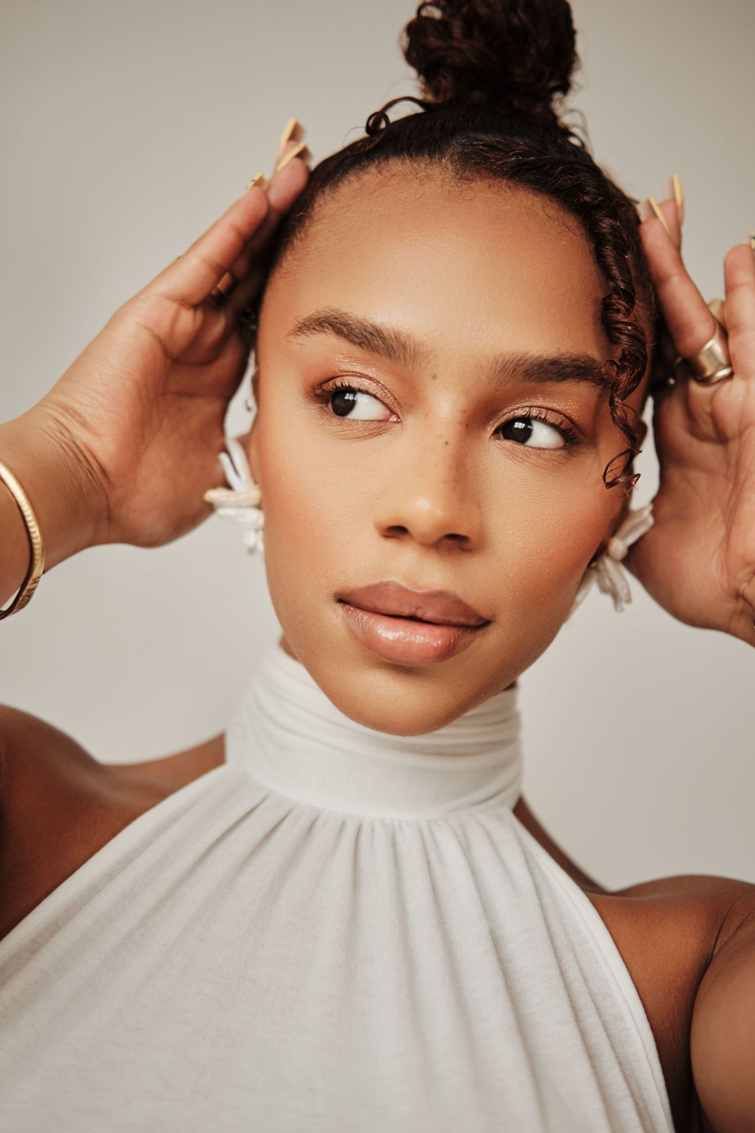 Young woman with hair bun wearing white turtleneck top, gold bracelets, hands raised to head, neutral background.