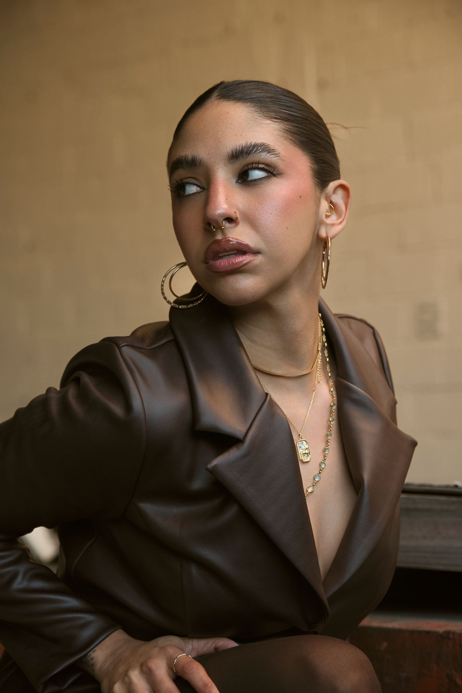 Professional portrait in brown leather blazer with gold jewelry against warm beige background.