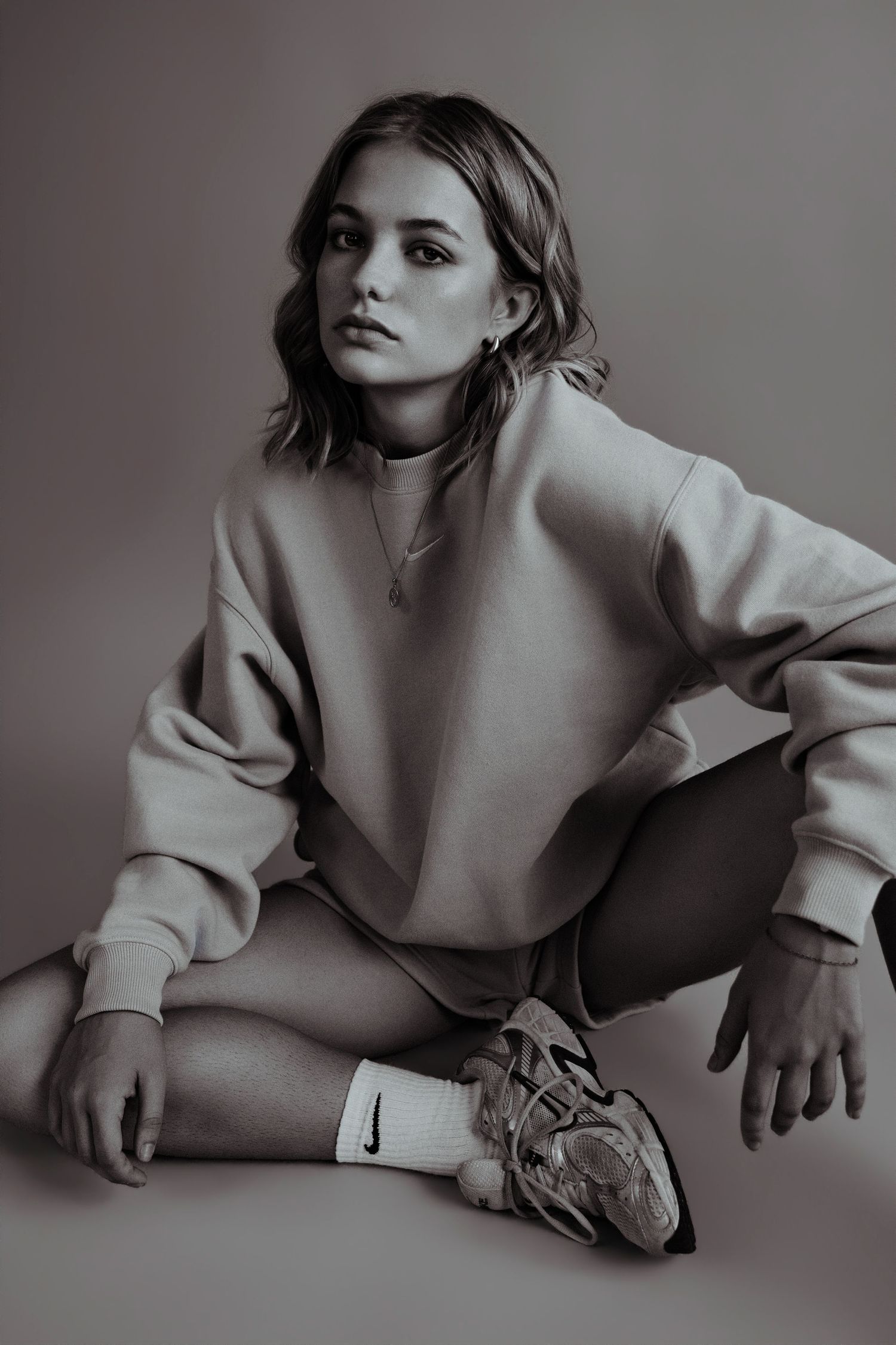 Young woman in oversized sweatshirt and Nike sneakers sits cross-legged on floor in moody monochrome studio photo.