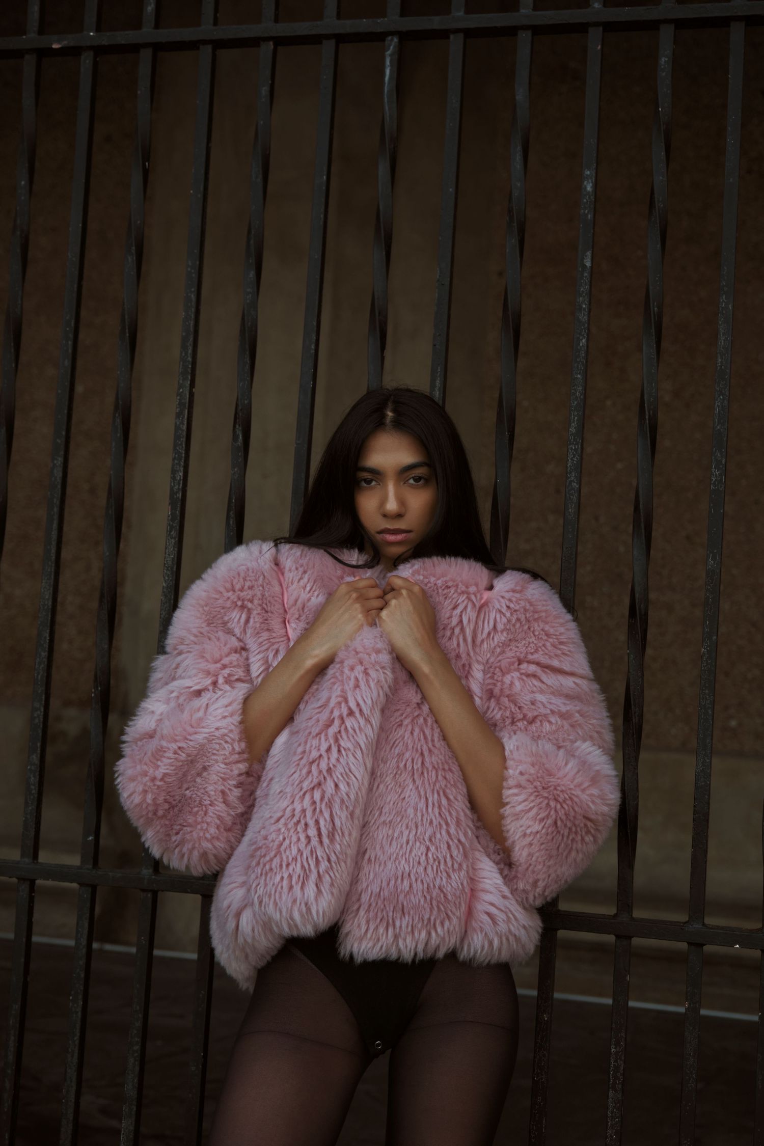 Dramatic fashion pose with pink faux fur jacket against dark metal bars and concrete backdrop.