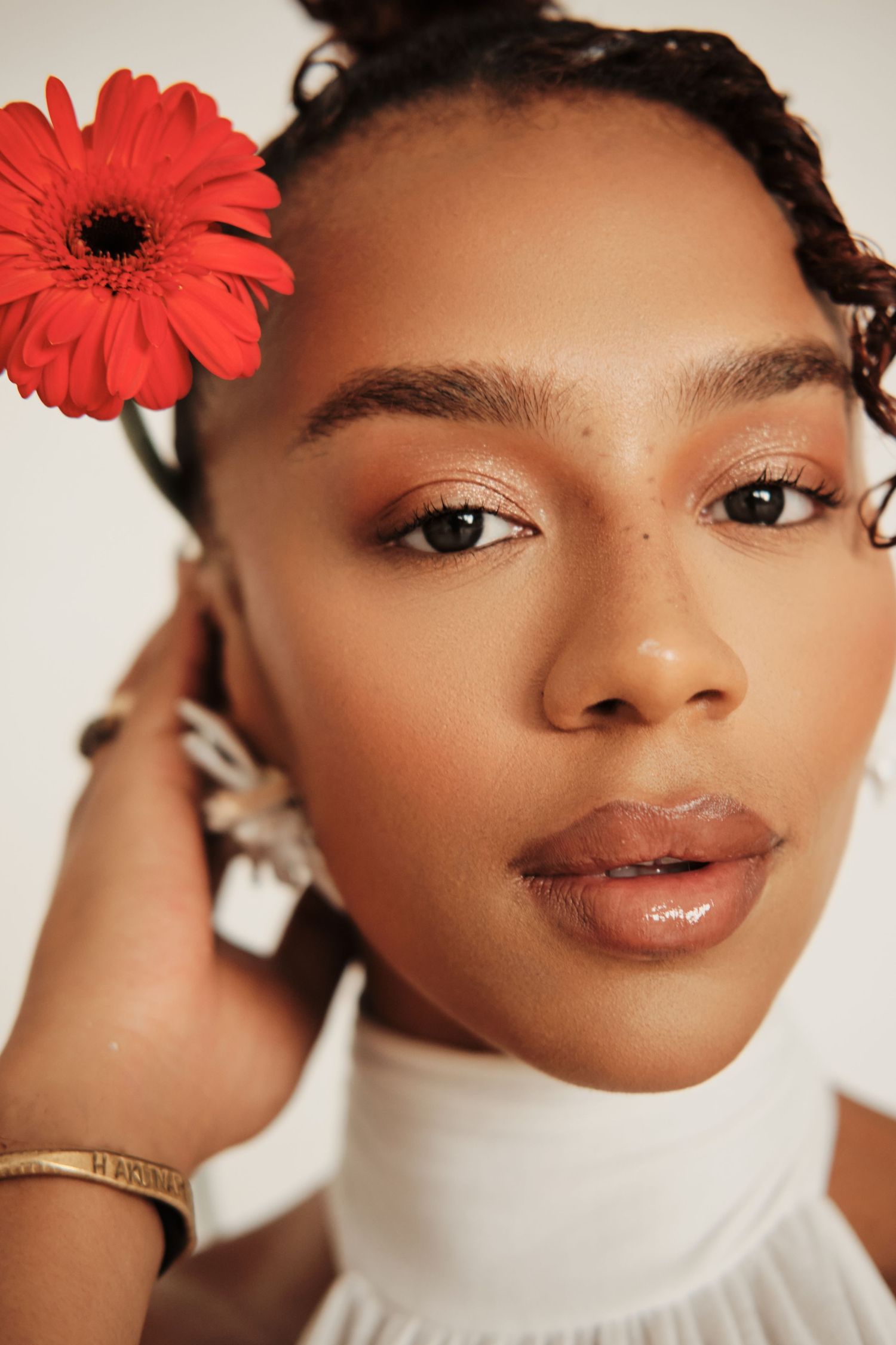Young Black woman with a red gerbera daisy in her hair, wearing a white turtleneck and gold bracelet, natural makeup look.