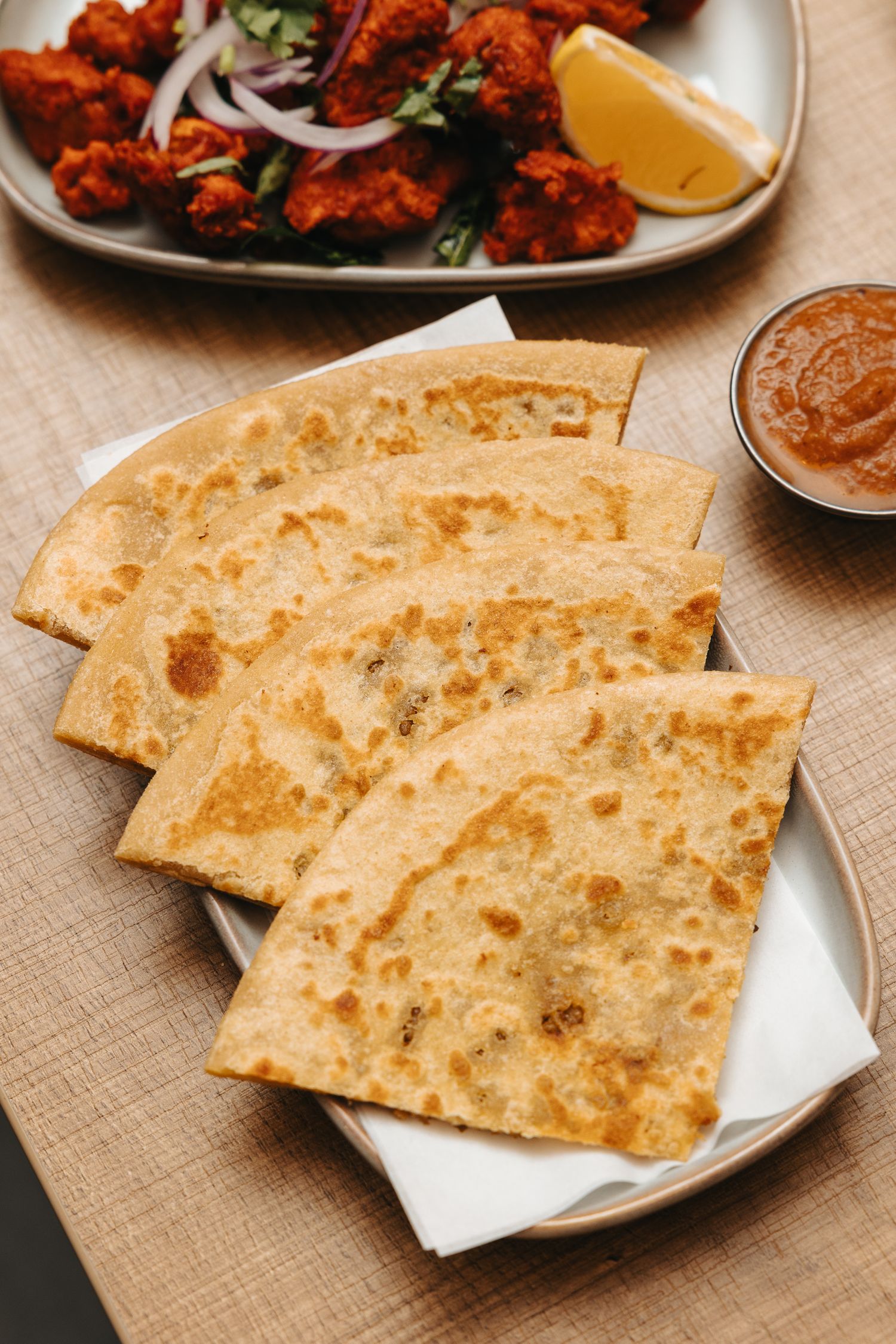 Golden-brown aloo paratha flatbreads served on a plate with dipping sauce, alongside spicy chicken tikka and lemon wedge.