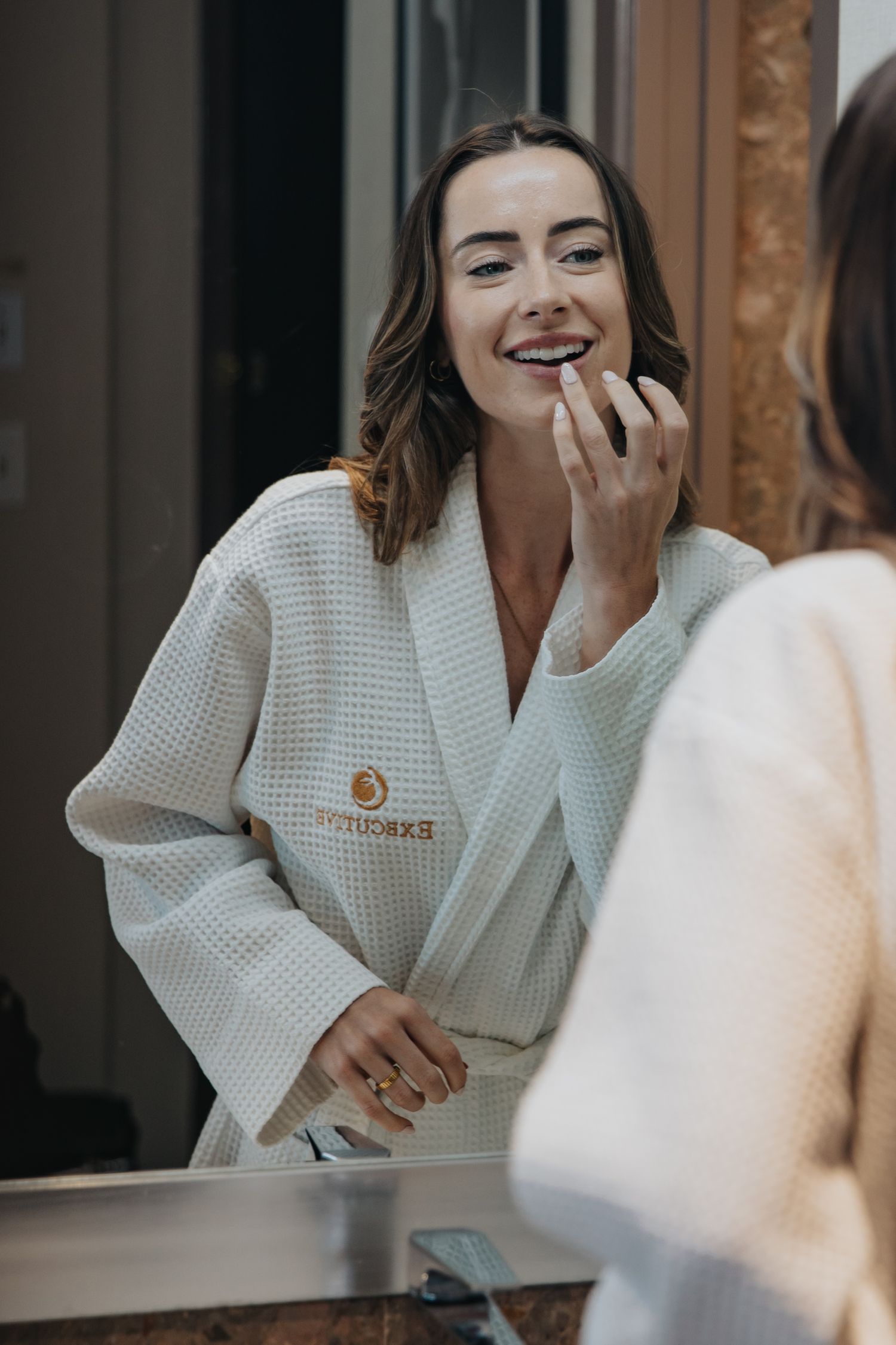 Smiling woman in a white waffle robe applying lipstick while looking in a bathroom mirror.