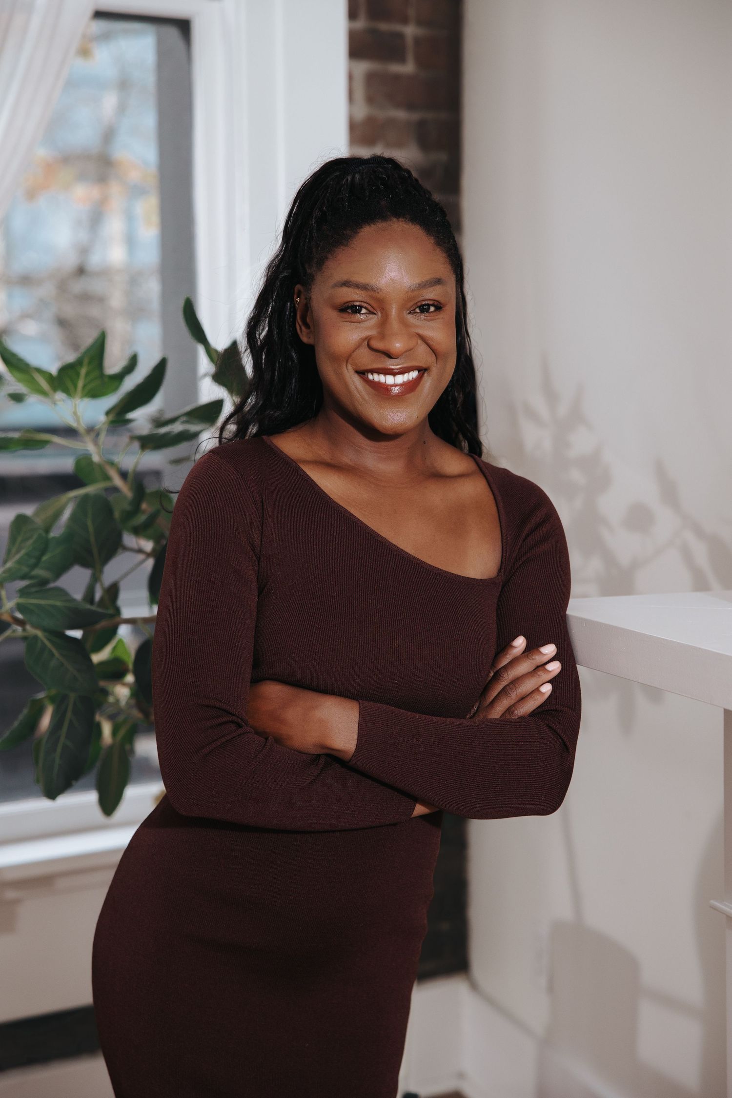 Professional portraits in brown sweater against white wall with window and plants.