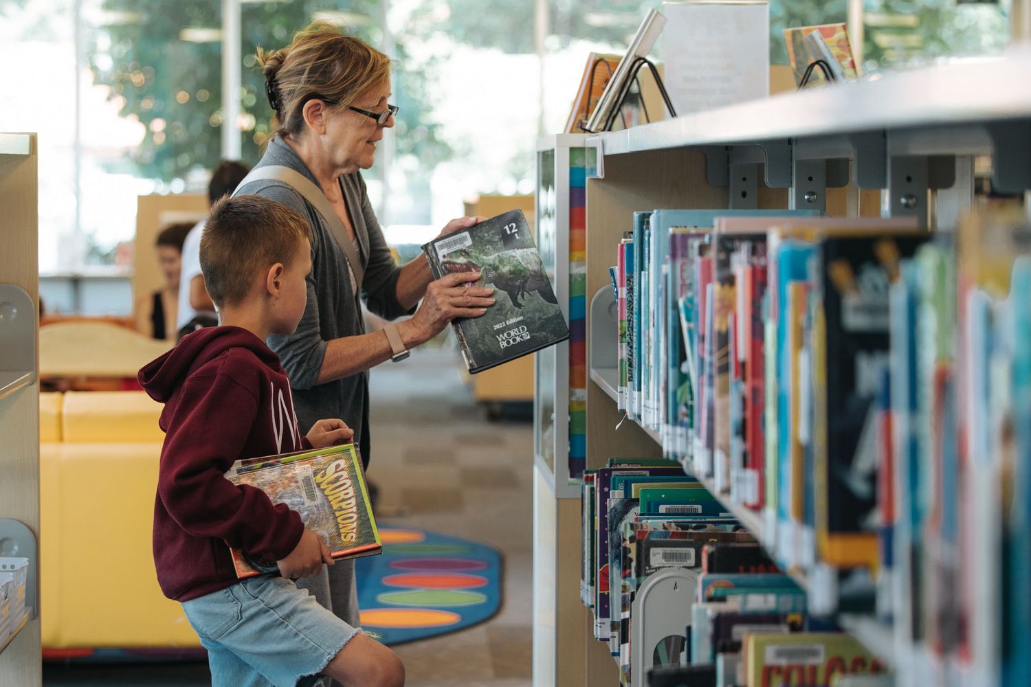 Adult and child browsing books together at library shelves.