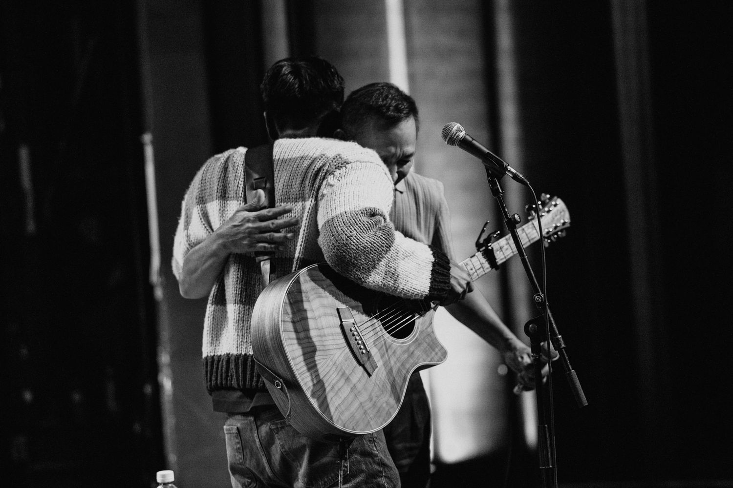 Two musicians sharing an embrace while playing guitars on stage in black and white photo.