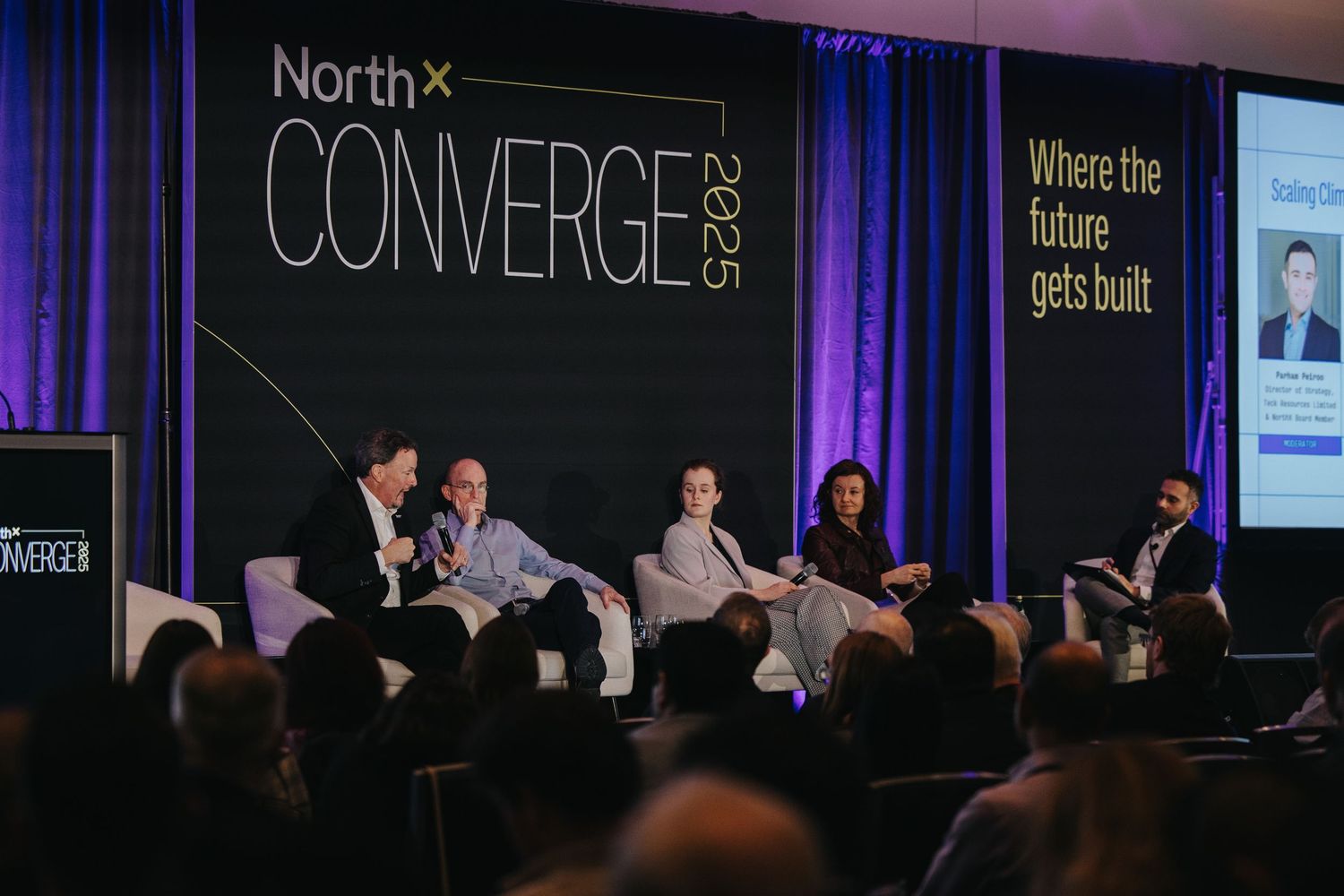 Business panel discussion on stage at North Converge conference with speakers seated in front of blue curtain backdrop.