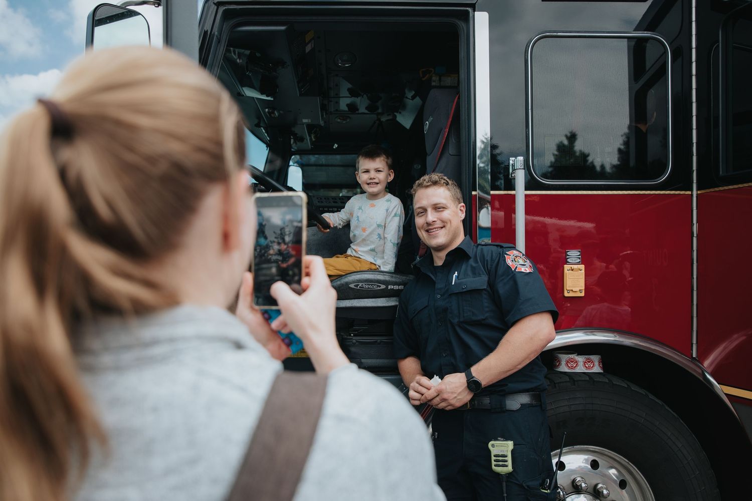 A firefighter in uniform stands next to a red fire truck while interacting with someone outside the frame.