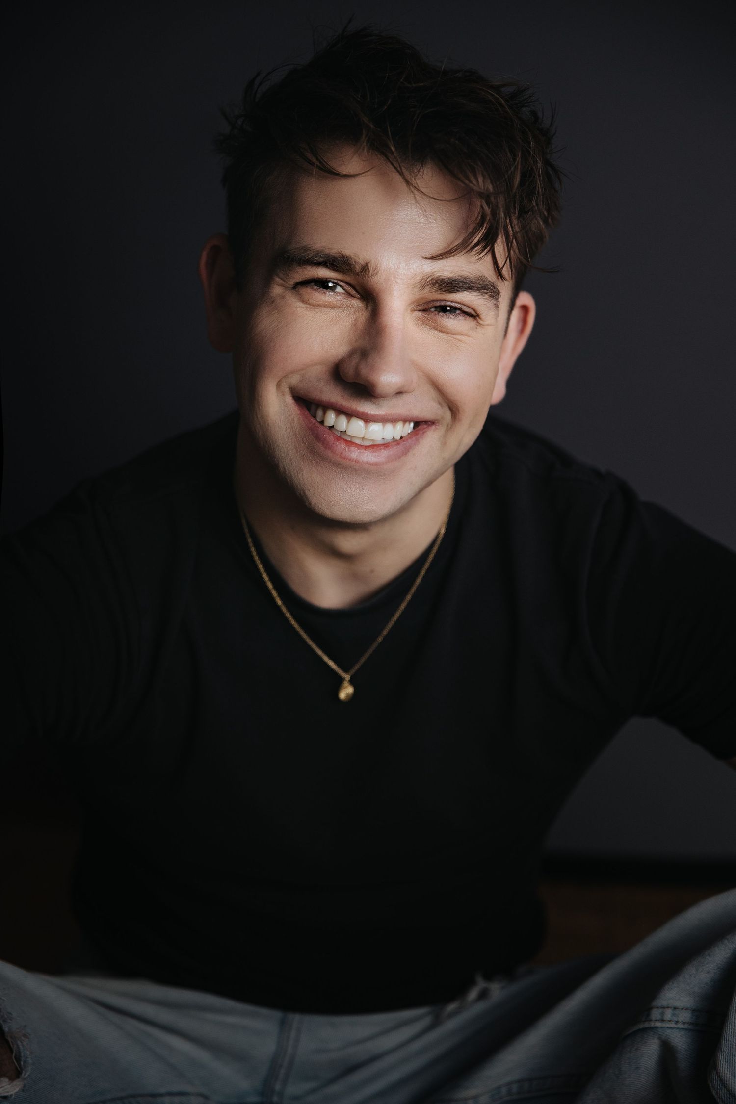 Studio portrait of person in black shirt with gold necklace against dark backdrop.