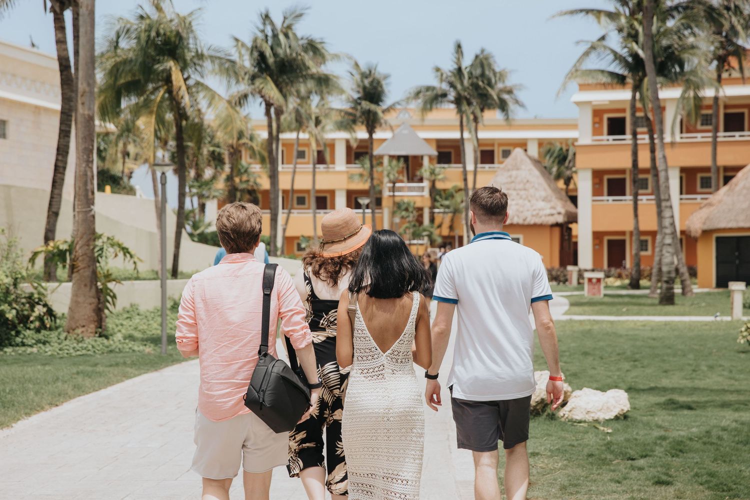 People walking along a tropical resort pathway lined with palm trees and peach-colored buildings.