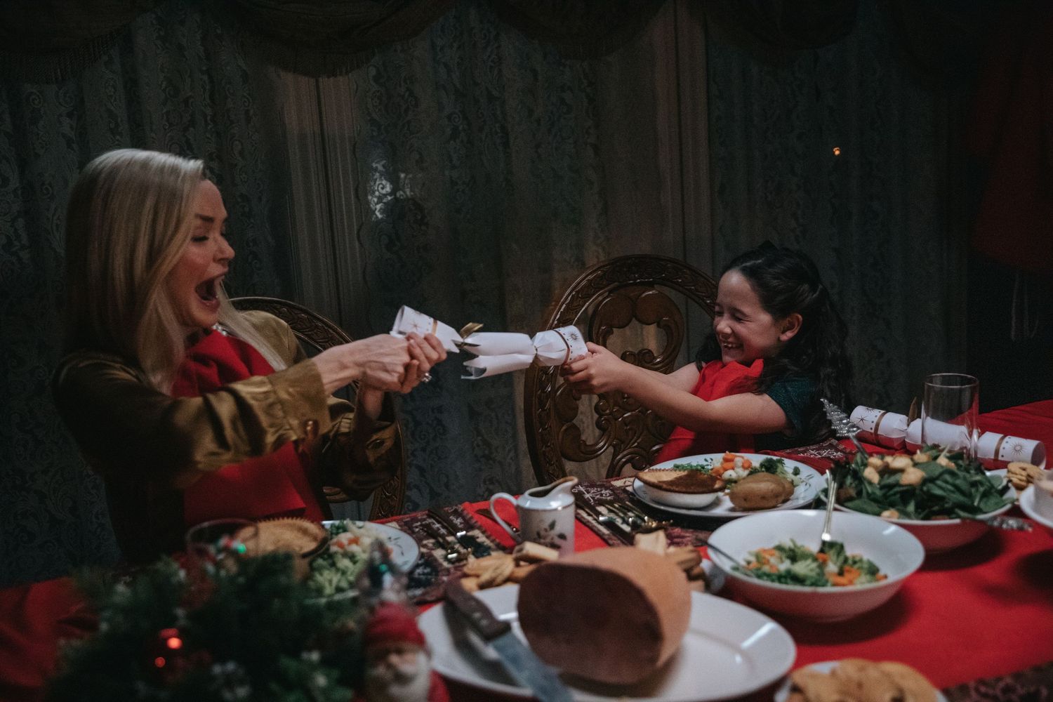 Two women in festive red outfits pulling a Christmas cracker at a holiday dinner table with decorations.