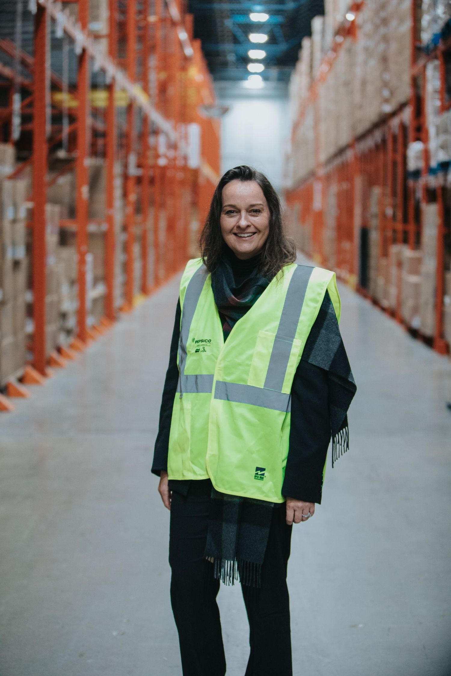 Woman in high-visibility vest smiling in a large warehouse aisle with orange shelving racks.