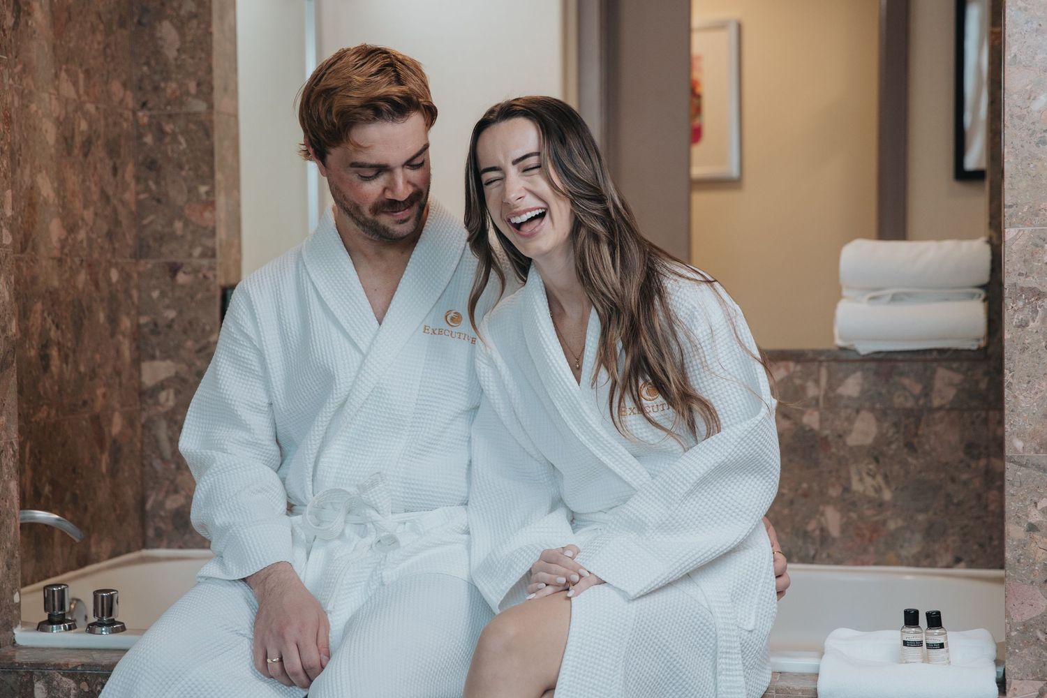 Couple laughing in white spa robes seated together in a luxury hotel bathroom spa area.