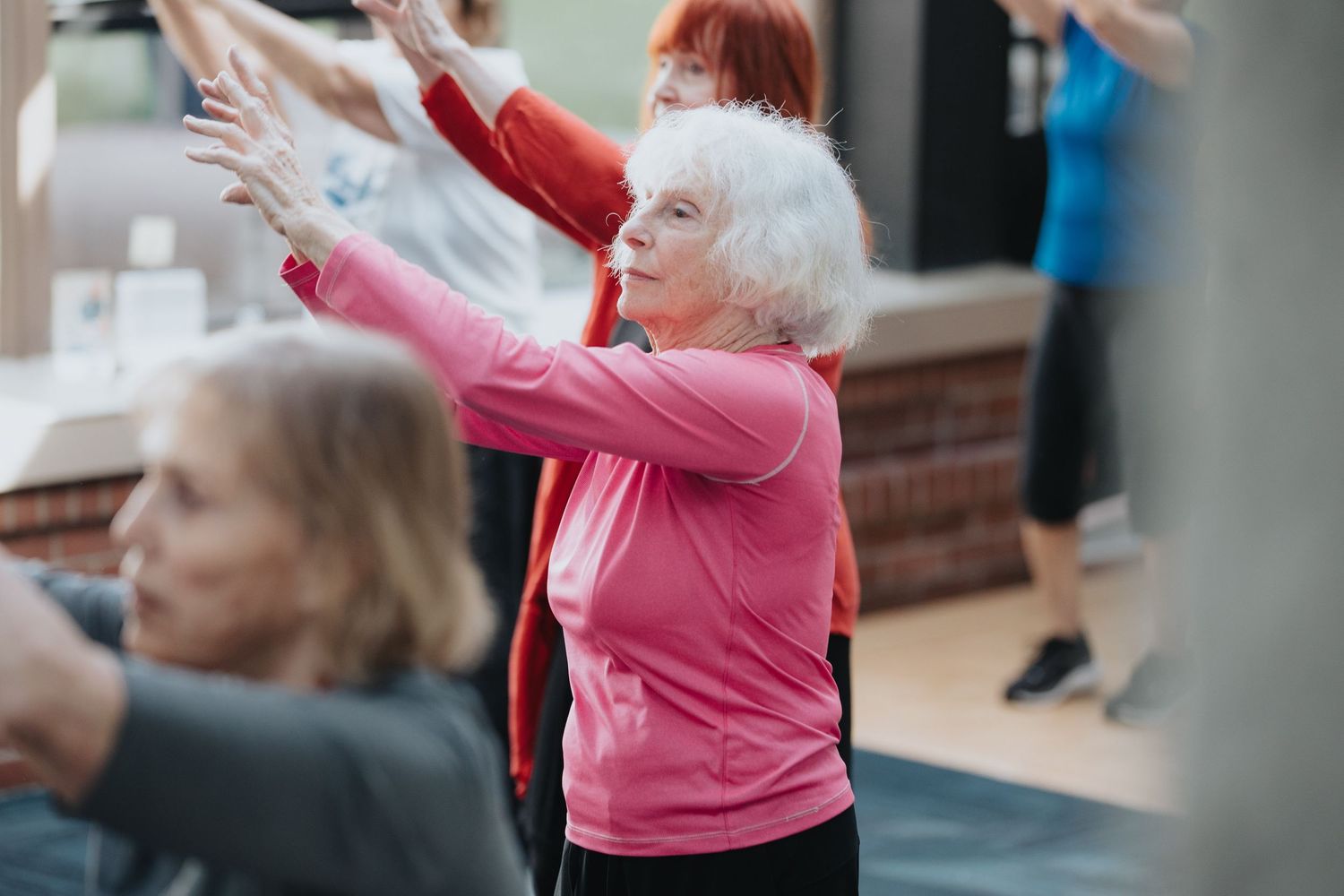 Elderly woman in pink top stretches arms during a group fitness class with other seniors in a bright studio.