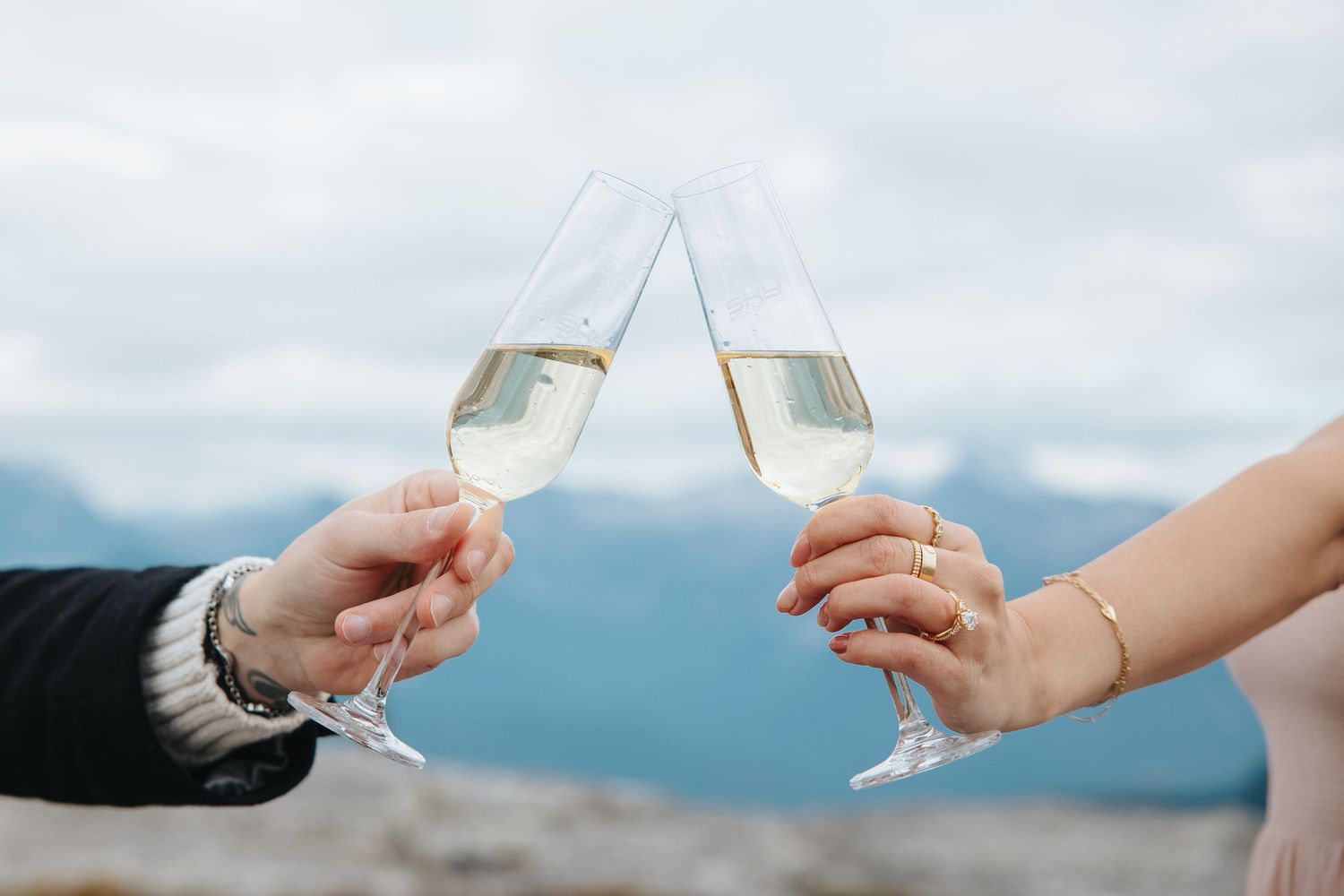 Two hands toasting champagne glasses against a scenic mountain backdrop.