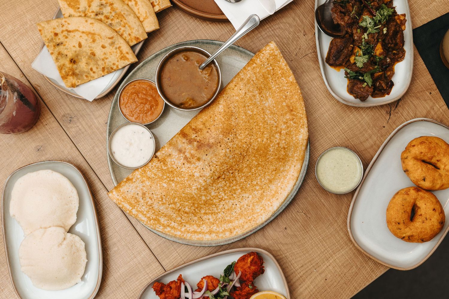 A spread of Indian dishes including a crispy dosa, idli, chutneys, paratha, and spiced meats on a wooden table.