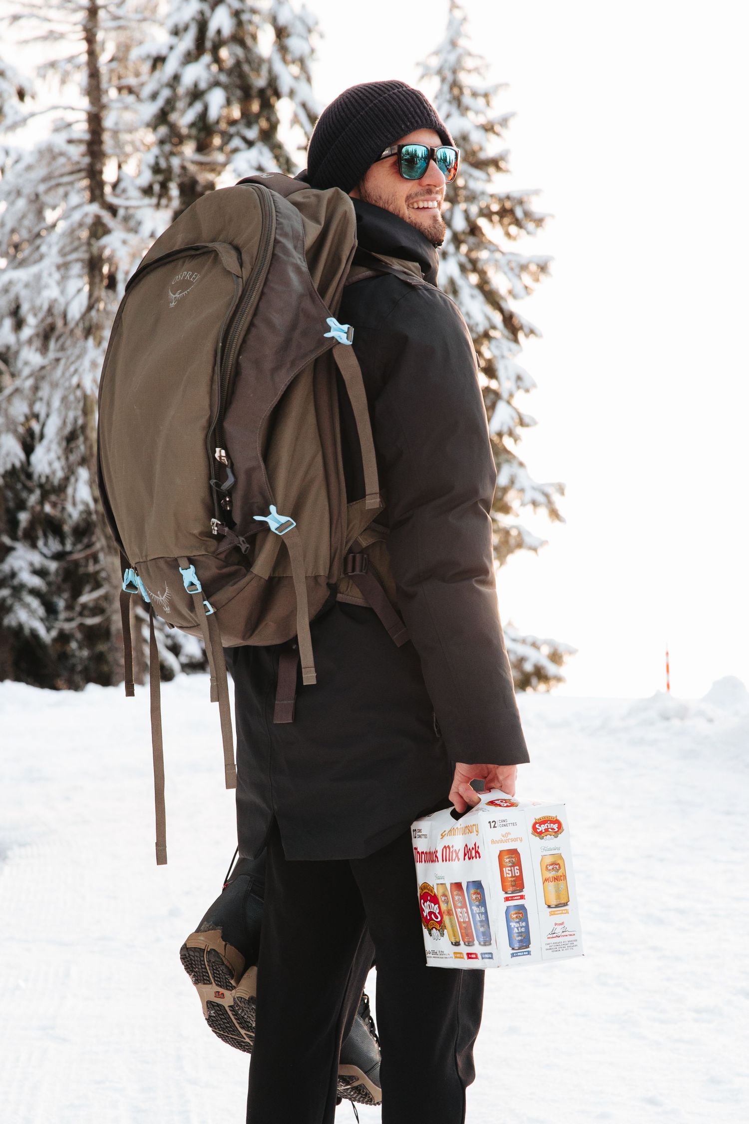 Hiker with backpack holding beverages while walking through snow-covered woods.