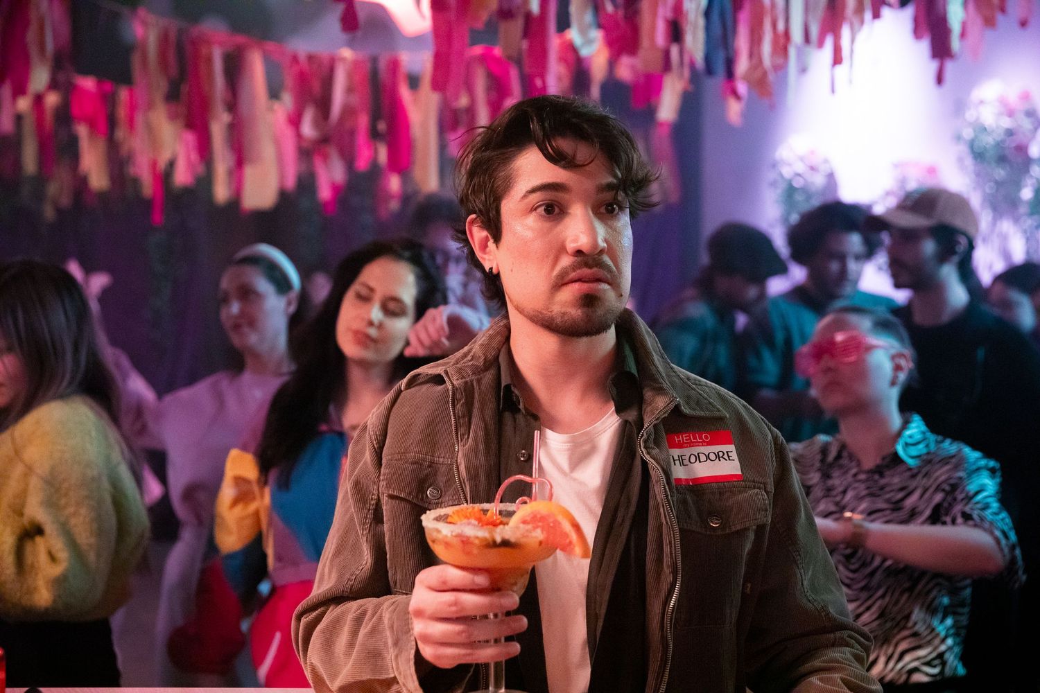 Young man holding an orange cocktail at a colorful party with pink decorations and crowd in background.