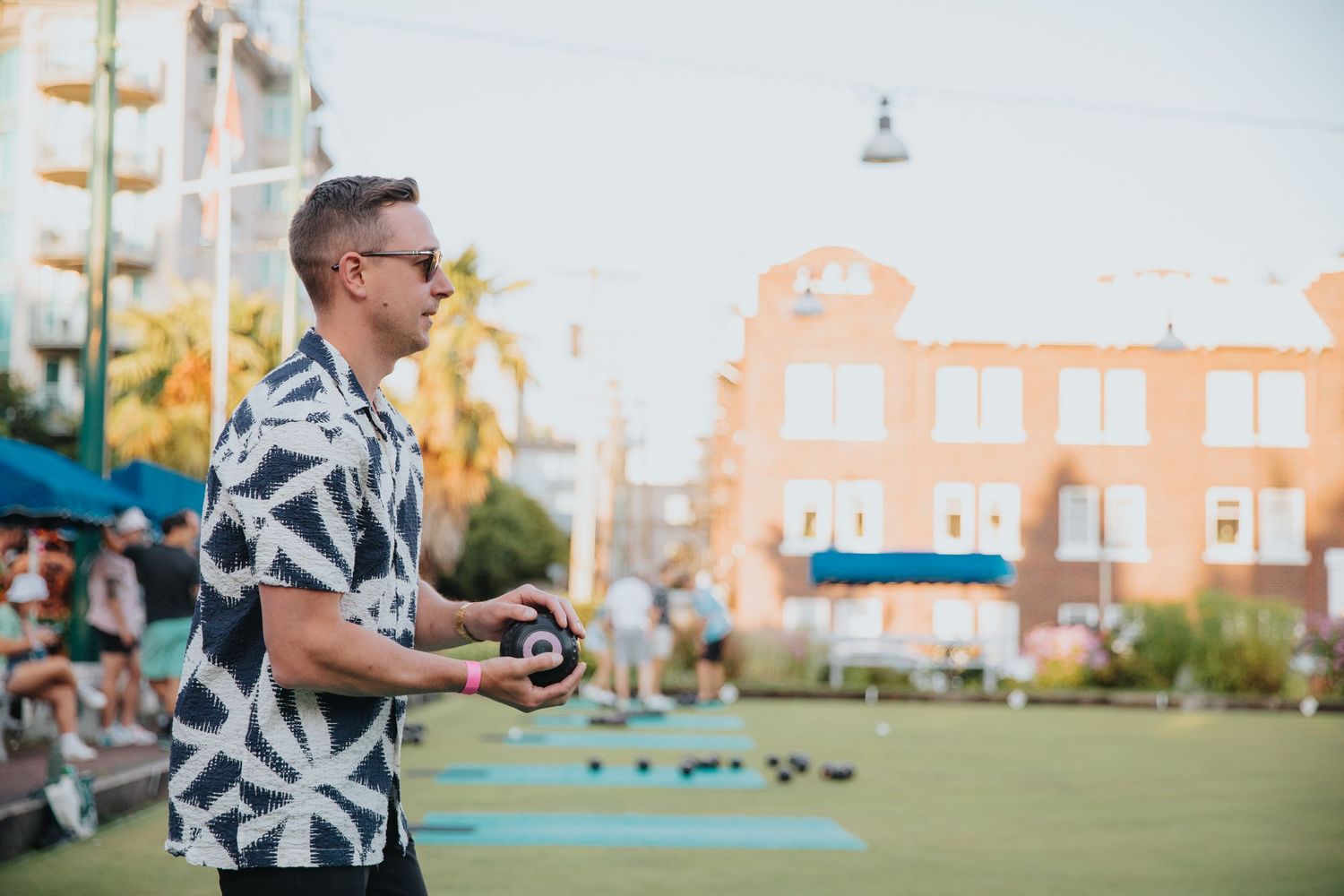A man in a patterned shirt plays bocce ball on an outdoor green court in an urban setting.