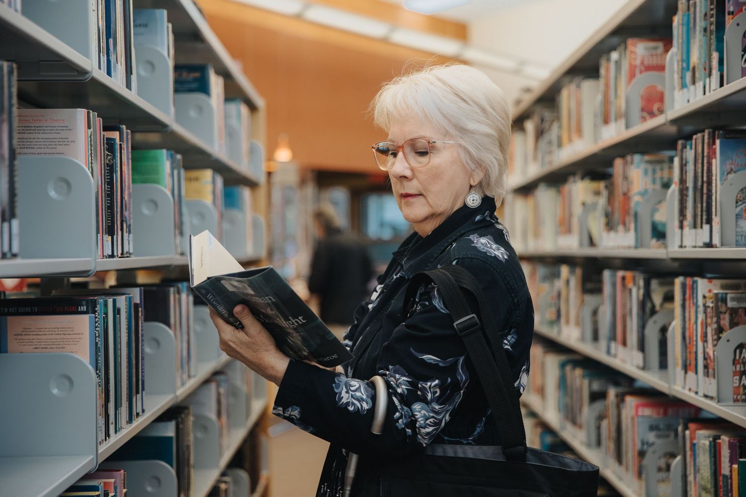 Person browsing books among library shelves in dark clothing.