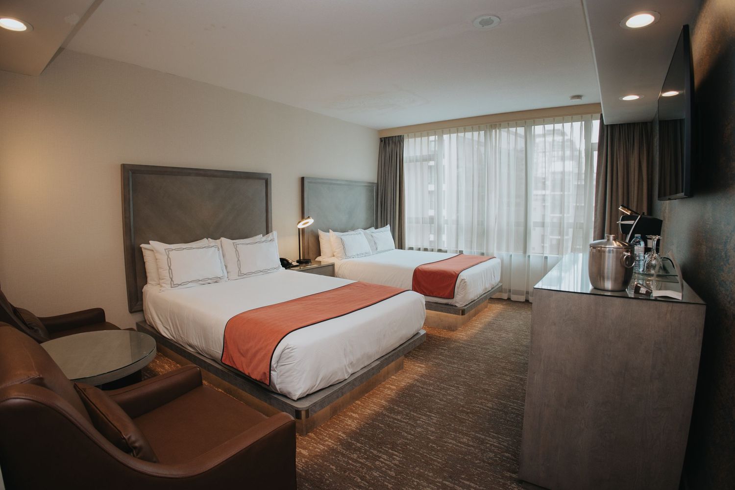 Spacious hotel room with two queen beds, warm lighting, leather chair, and city view window.