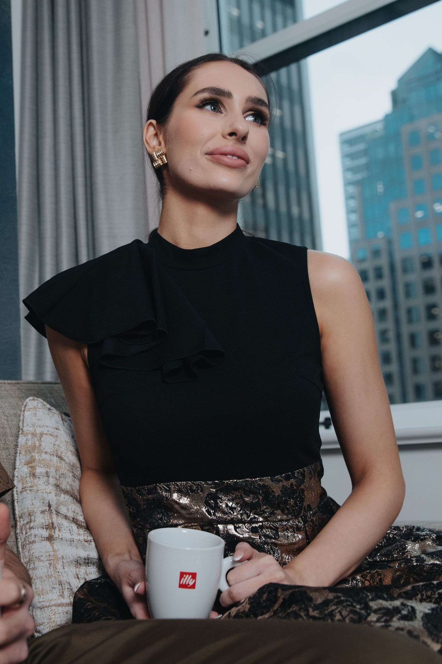 Woman in black dress sitting by window with city skyline view.