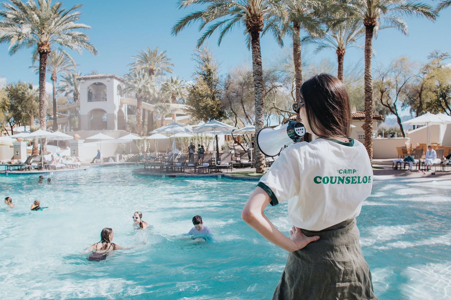 People splashing and playing in a resort swimming pool surrounded by palm trees.