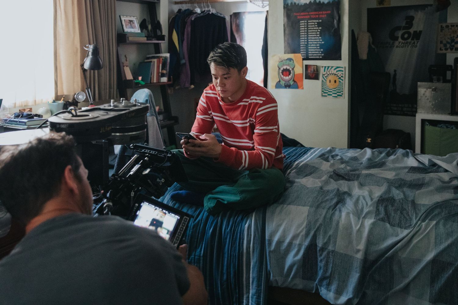 Young man in striped shirt sits on bed using phone while being filmed in a cluttered dorm room.