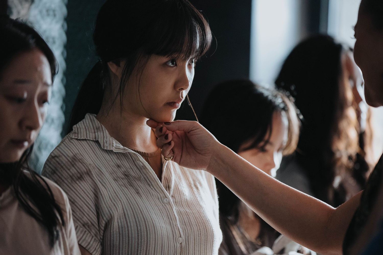 A young Asian woman in a white shirt gets makeup applied backstage surrounded by others.