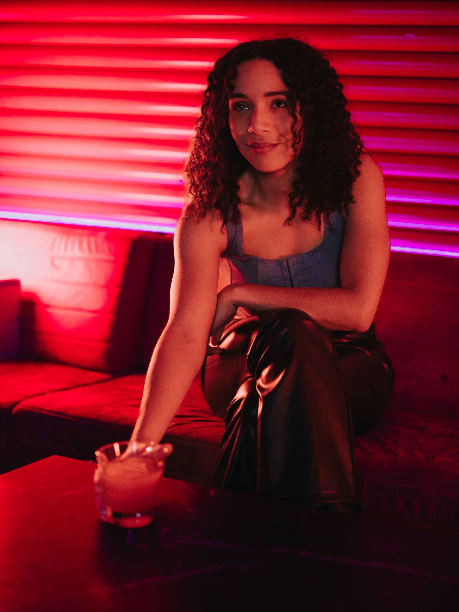 Young woman with curly hair sitting on a couch in a nightclub with red neon lighting holding a cocktail.
