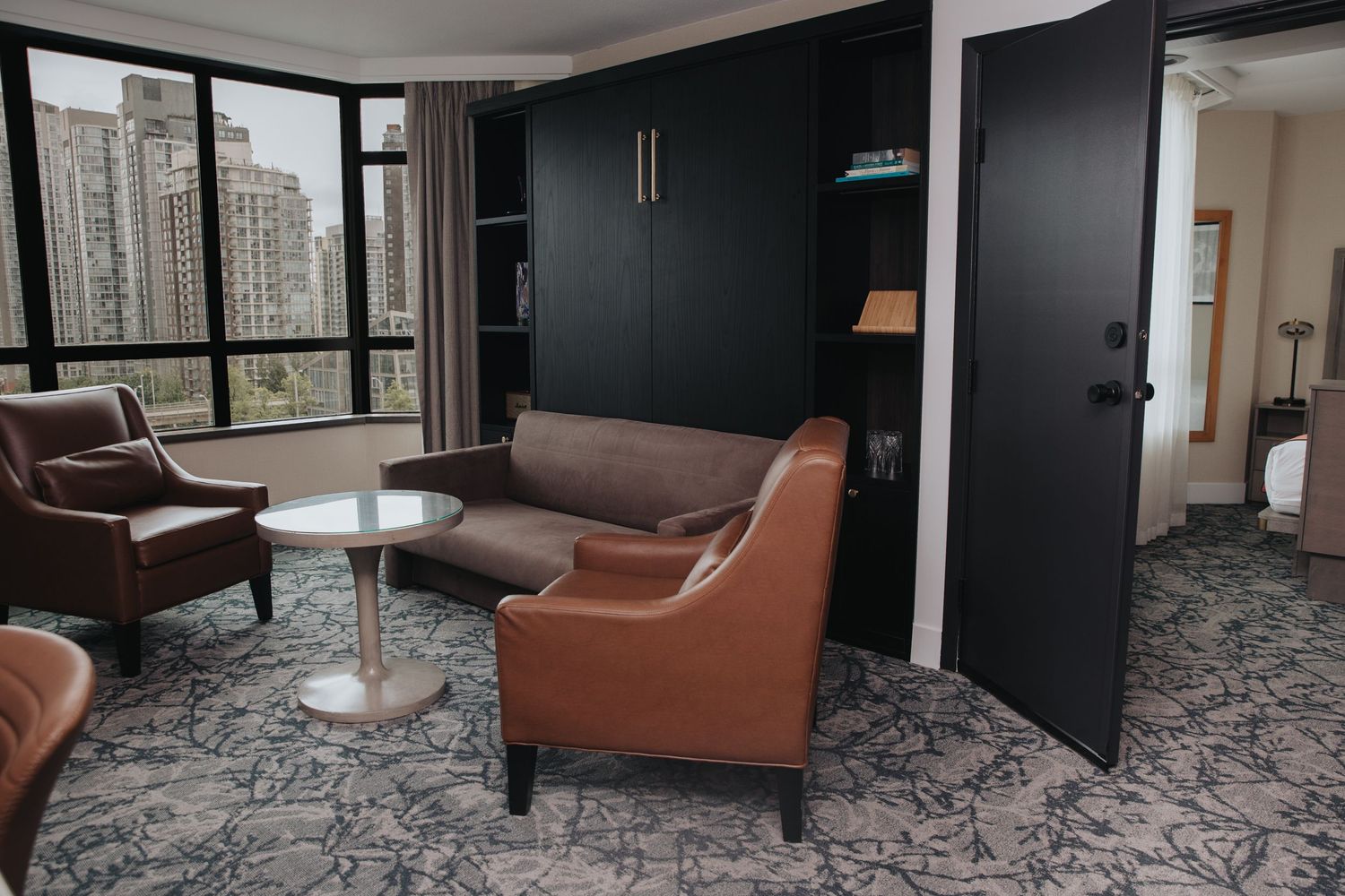 Modern hotel suite living area with leather chairs, sofa, and floor-to-ceiling city view windows.