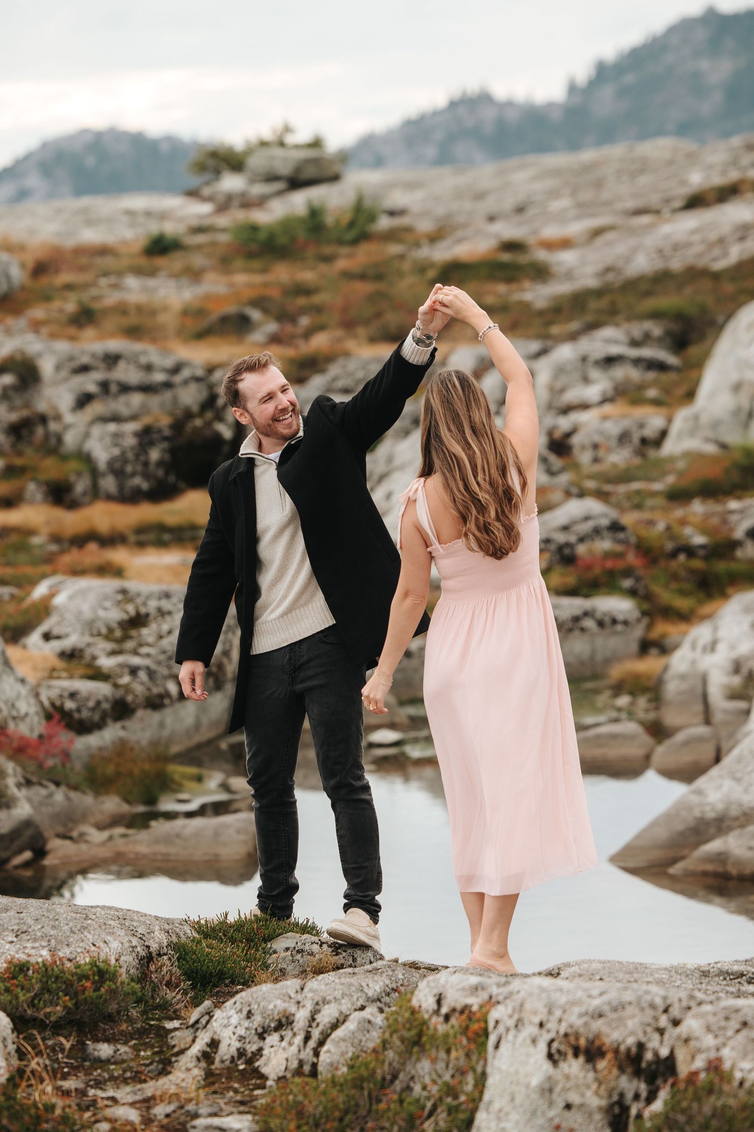 A couple dancing on rocky terrain with mountains and water in the background.