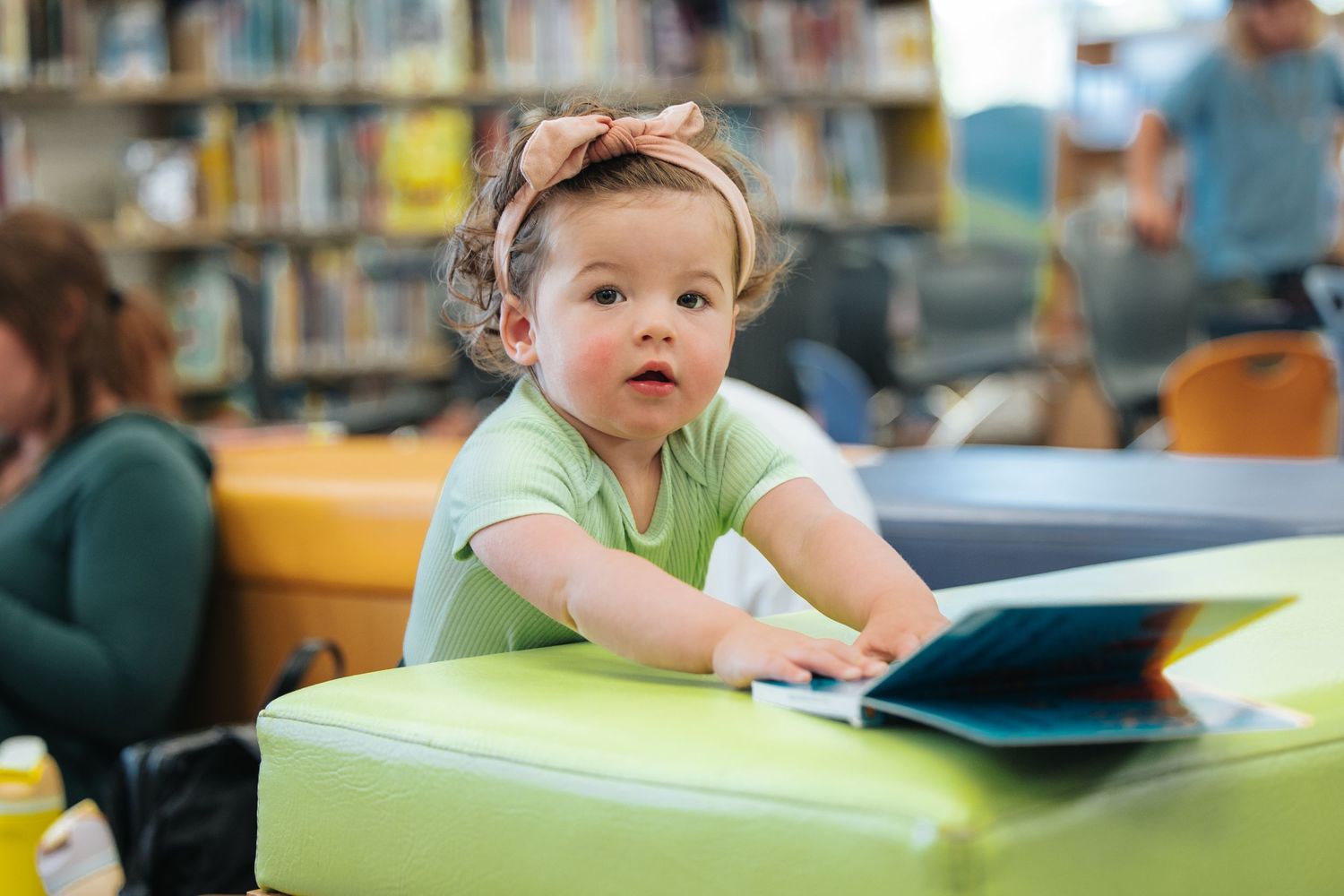 Young child in a green shirt using a tablet device at a library table.