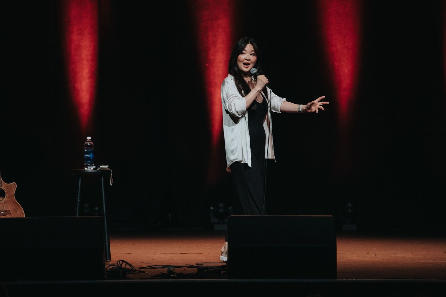 Female comedian performs on stage with red dramatic lighting and guitar visible in background.