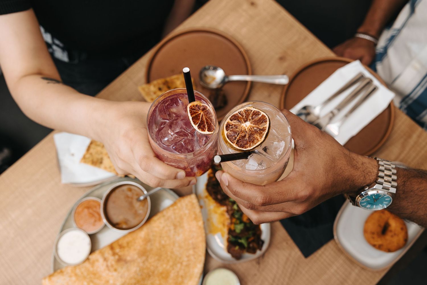 Two people toasting cocktails garnished with dried orange slices over a table of food at a restaurant.