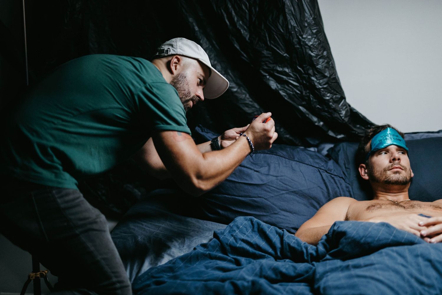 A man in a green shirt and cap tends to another man lying in bed inside a dark tent.