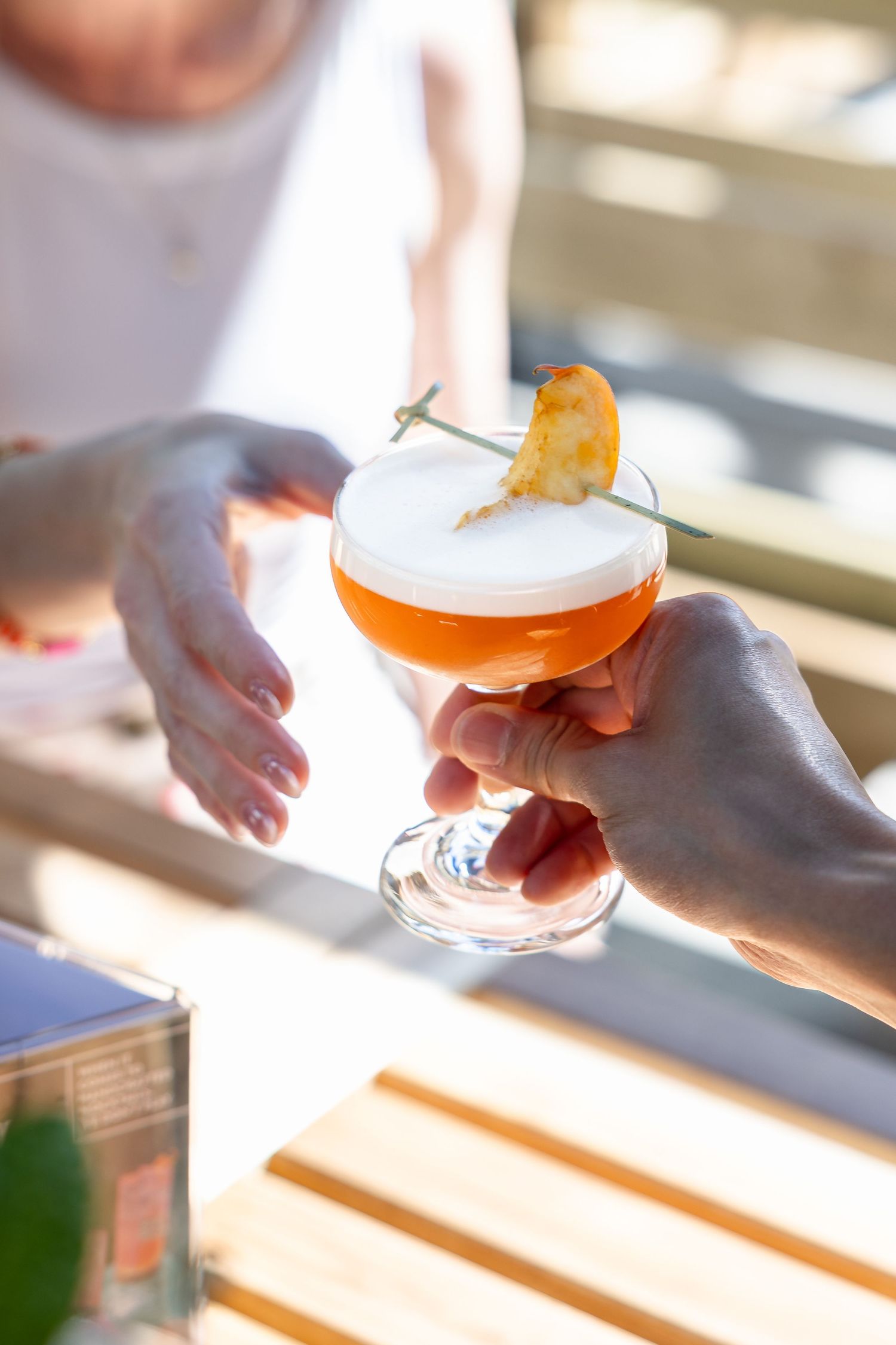 A hand holding an orange cocktail topped with white foam and a citrus garnish on a sunny outdoor patio.