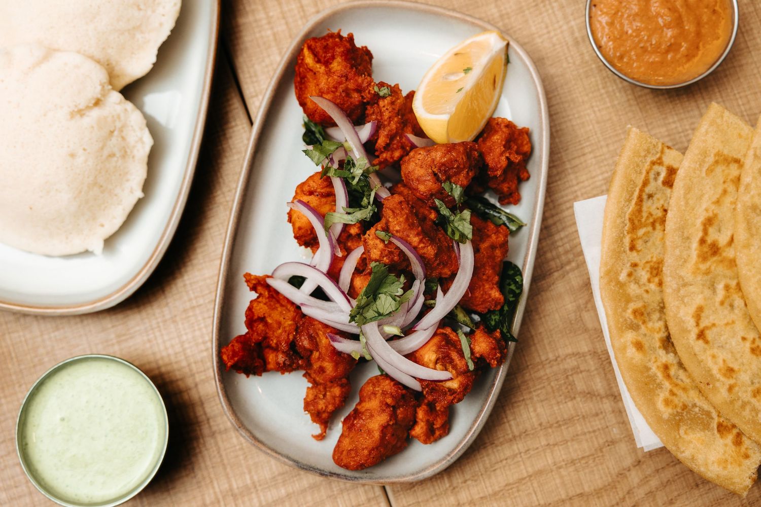 Spicy Indian chicken 65 served on a platter with onions, curry leaves, lemon, alongside idli, chutney, and paratha.