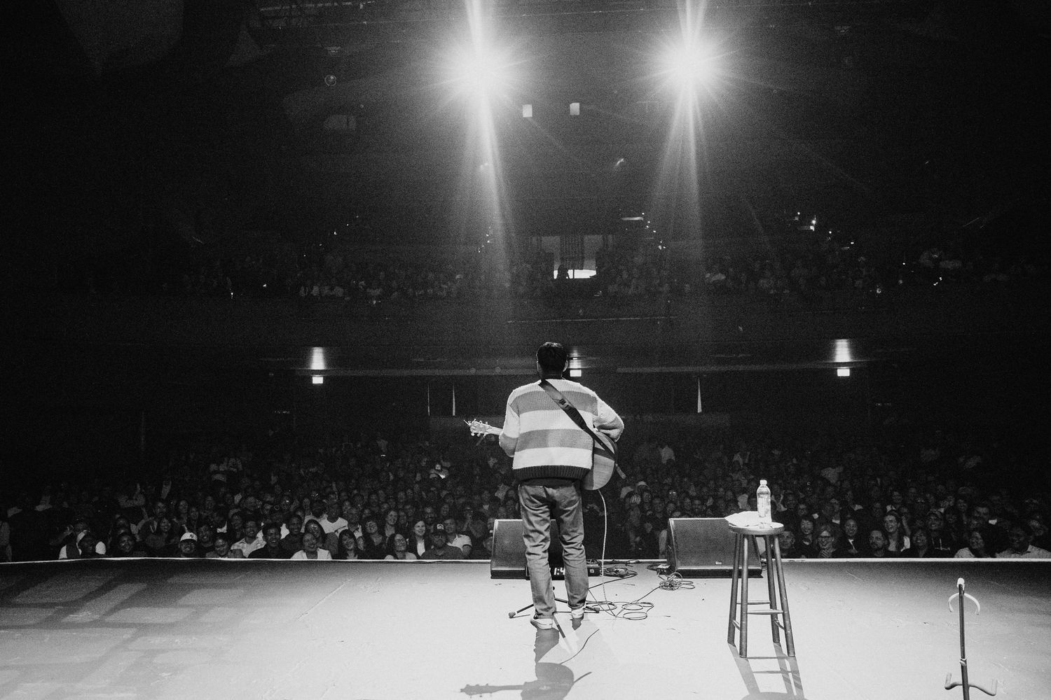 Performer on stage facing large arena crowd under bright spotlights in black and white photo.