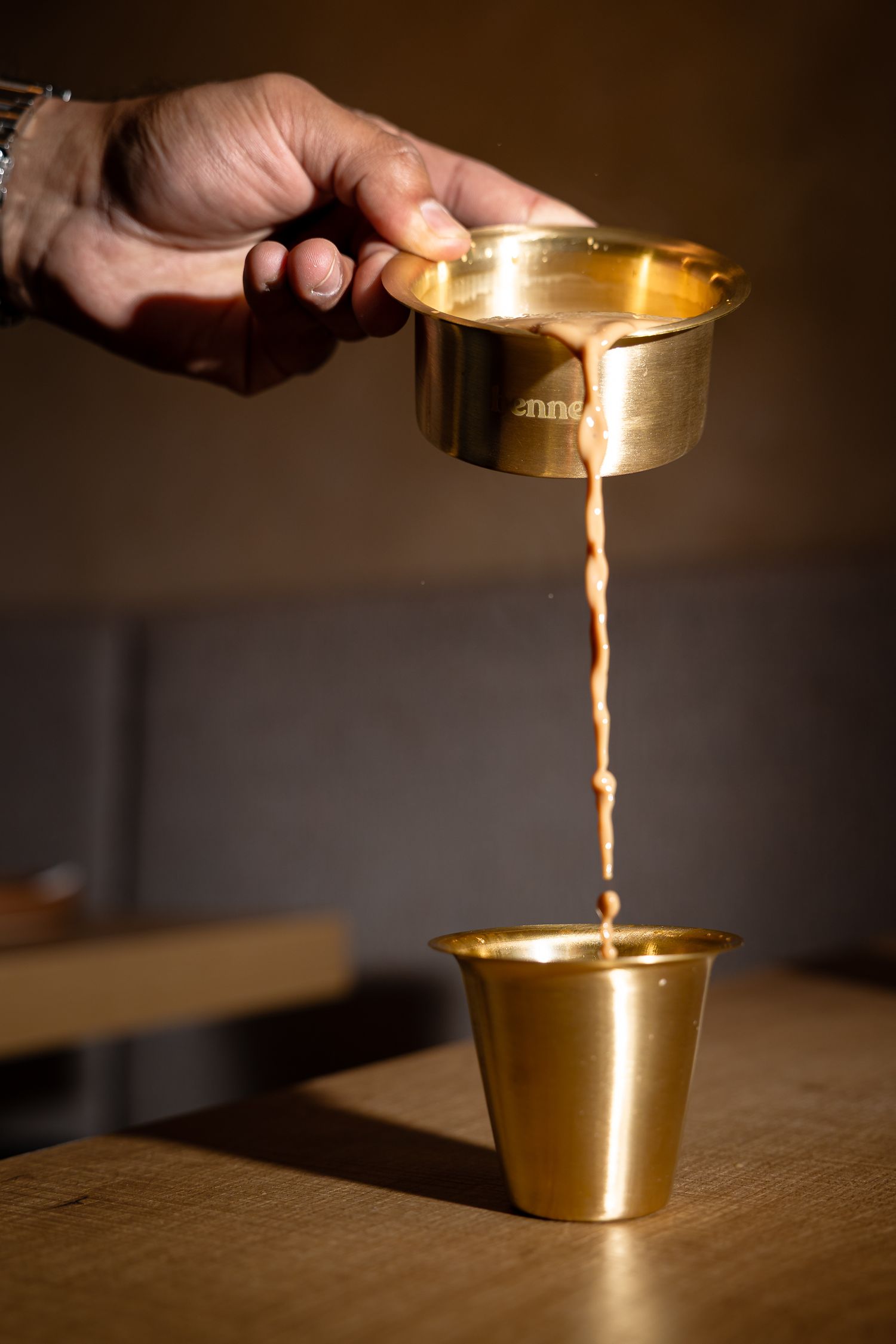 Hand pouring espresso from a gold portafilter into a golden cup on a wooden surface.