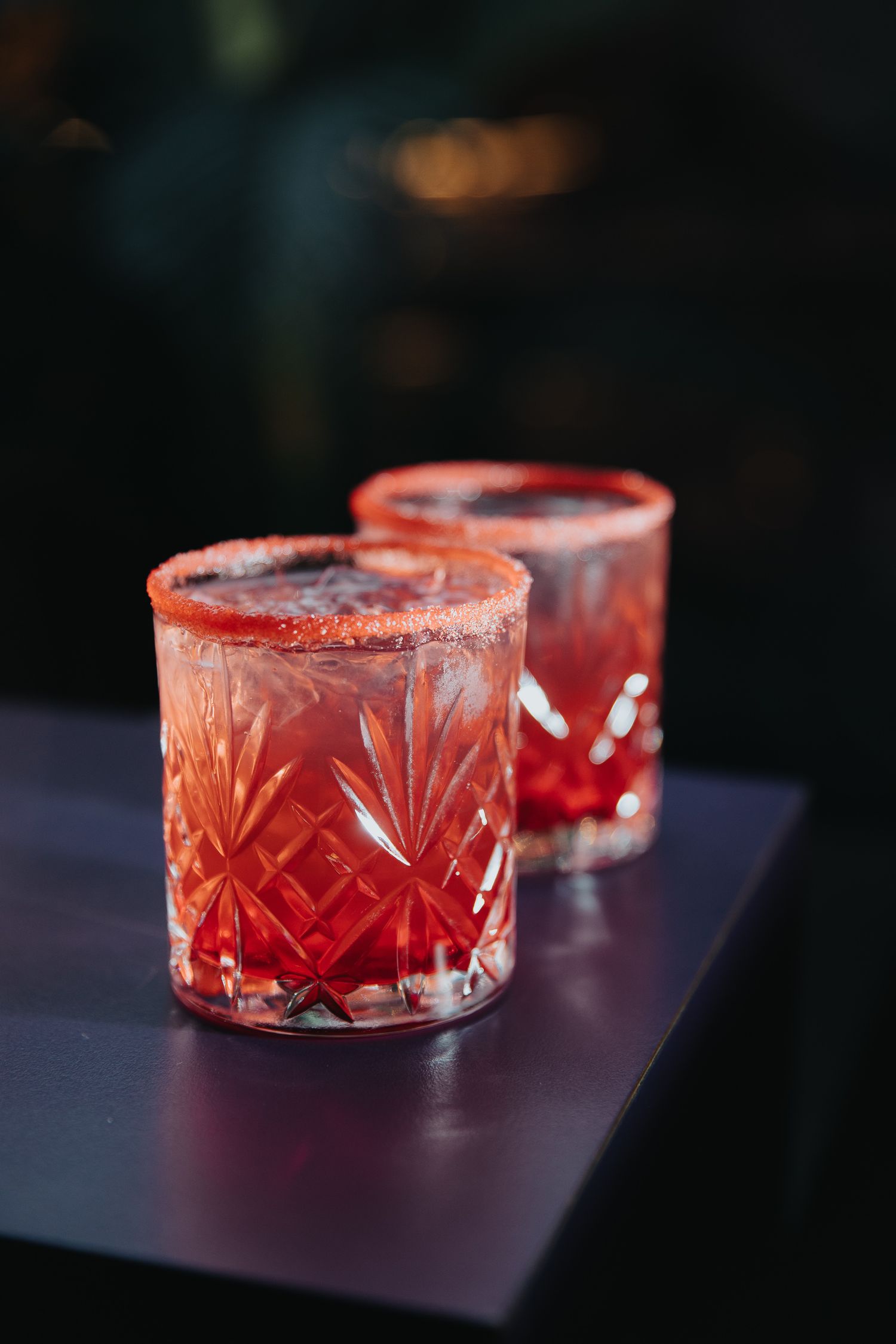 Crystal cocktail glasses with red drinks and decorative sugar rims on a dark surface.