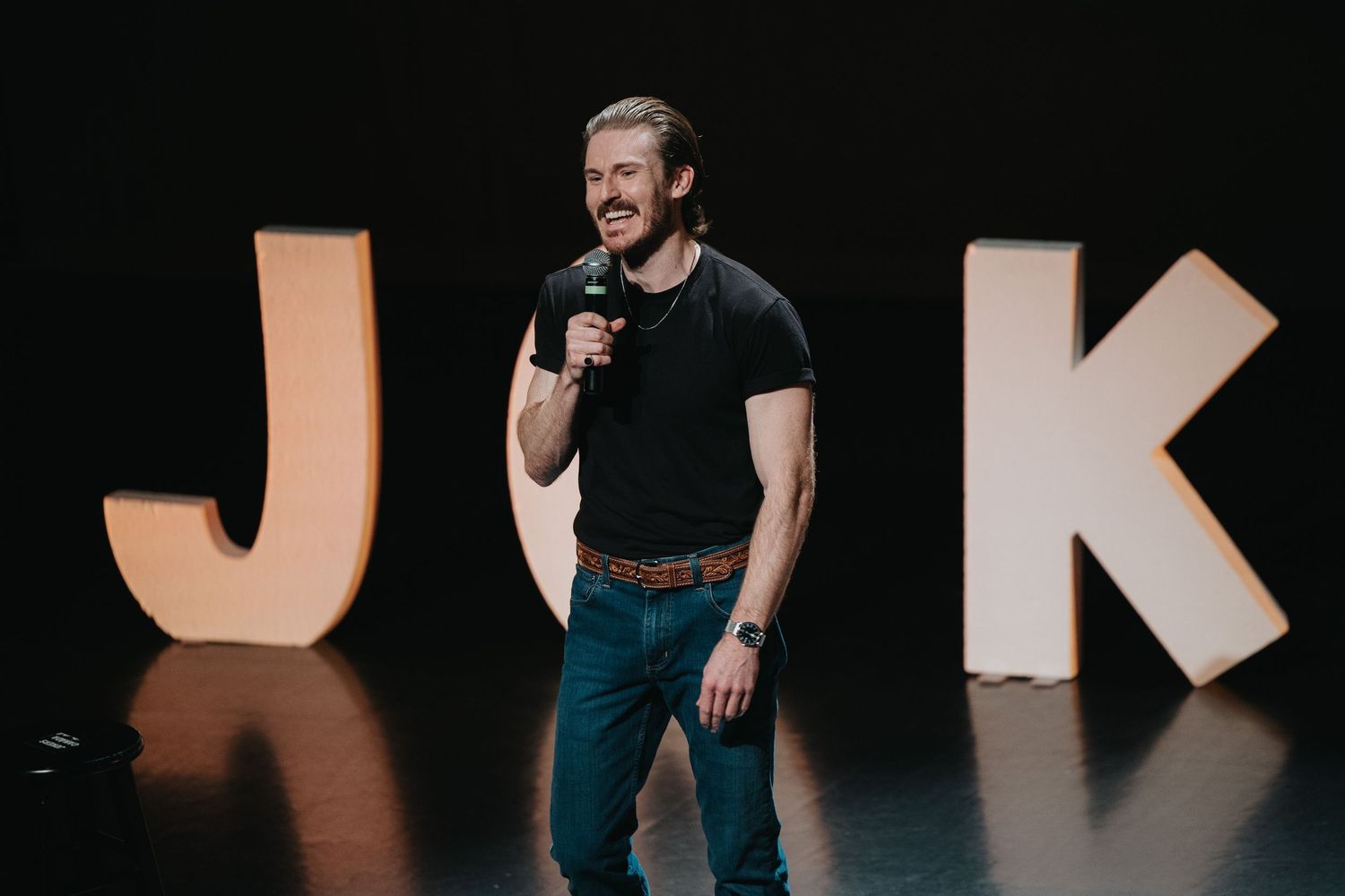 Man in black t-shirt performs stand-up comedy on stage with large illuminated J and K letters backdrop.