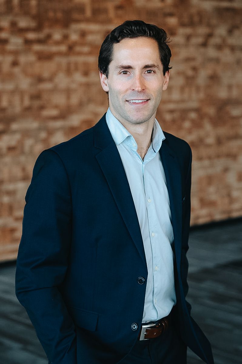 Professional headshot portrait against brick wall backdrop.