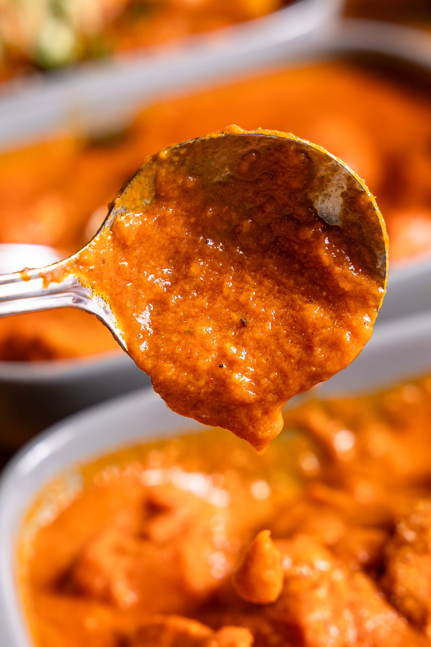 Close-up of a spoon holding rich, creamy orange curry sauce over a bowl of chicken curry.